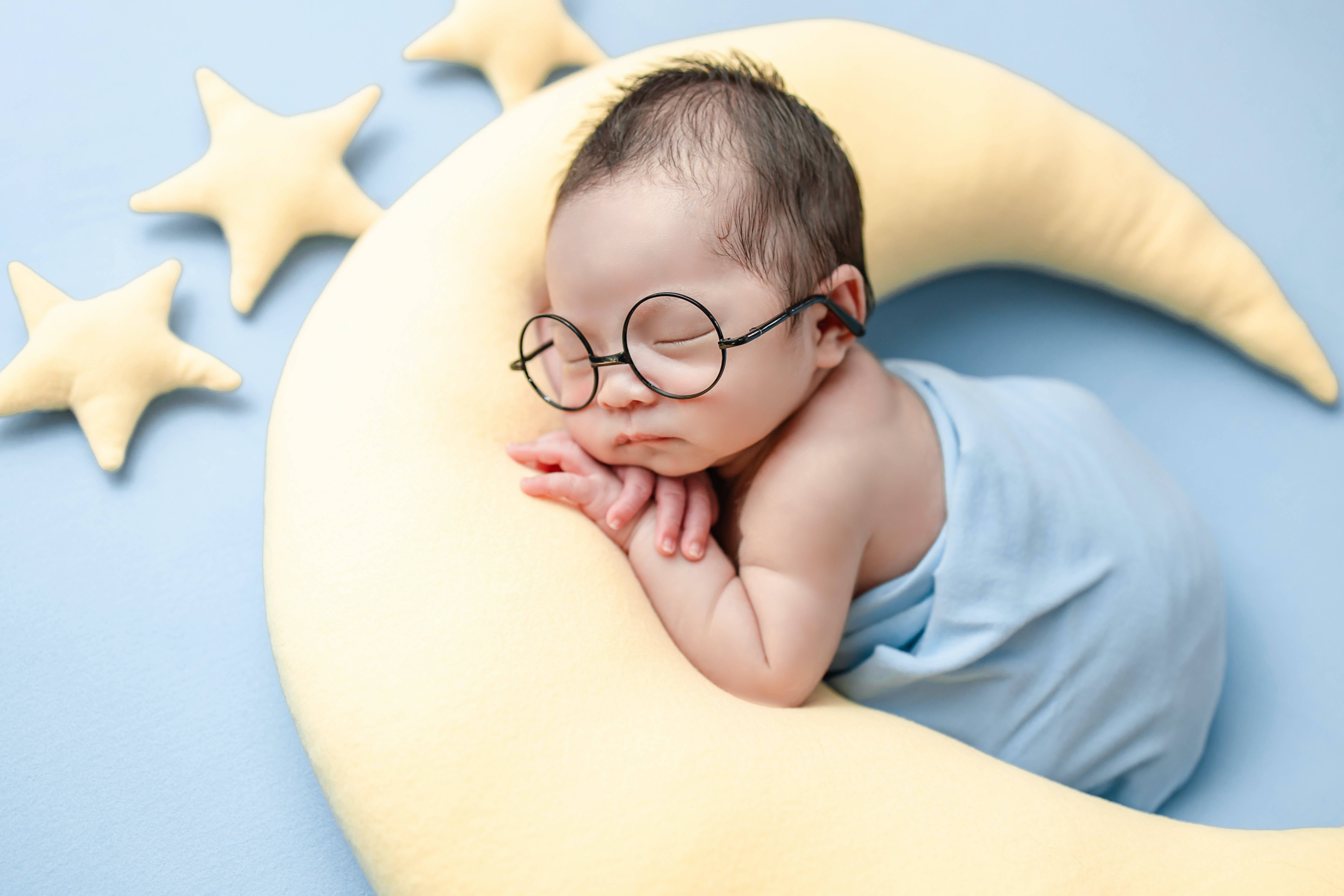 戴眼鏡的超可愛嬰兒 壁紙 — 一個戴著圓形眼鏡的寧靜新生嬰兒，在星星環繞的月牙枕頭上安然入睡。
