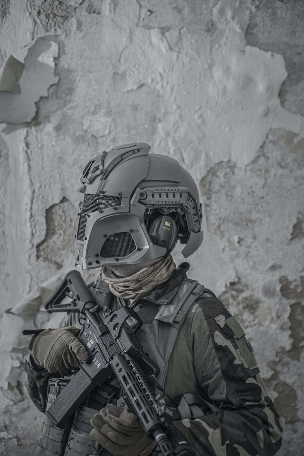 戴磨砂灰頭盔的軍人制服 壁紙 — 一名身穿全套戰術裝備和頭盔的士兵，靠著風化的混凝土牆，手持突擊步槍保持警覺。