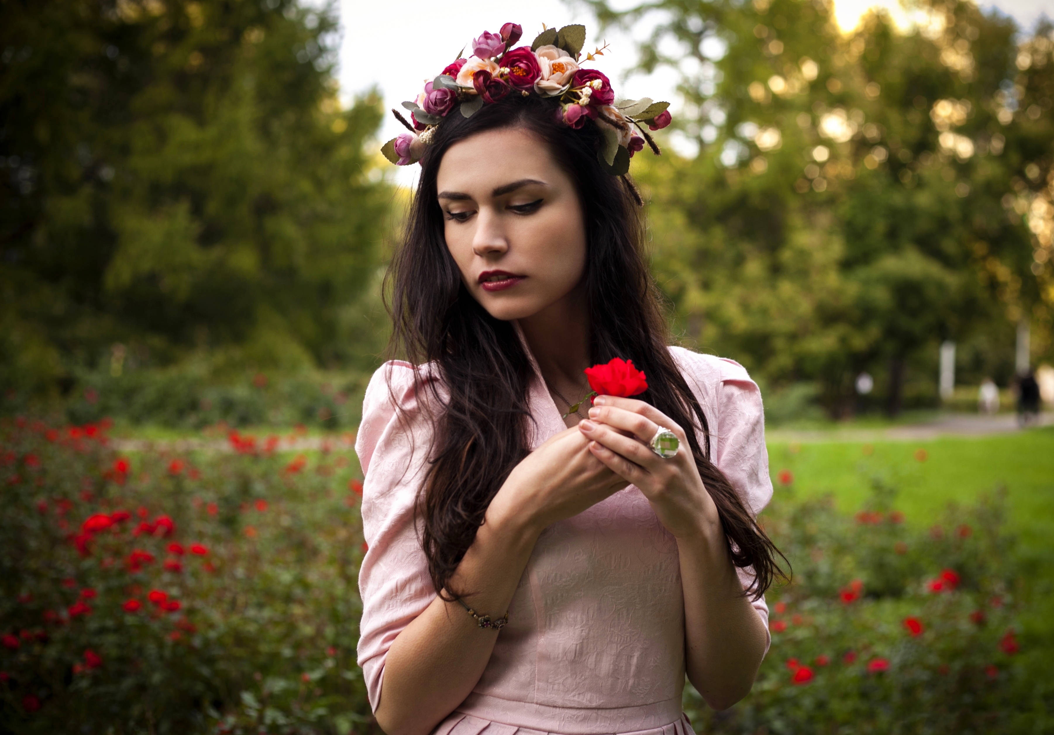戴粉紅花朵冠冕的可愛女孩 壁紙 — 一位身穿粉紅色連衣裙、頭戴花冠的女性在陽光普照、開滿玫瑰的花園中輕輕地拿著一朵紅玫瑰。
