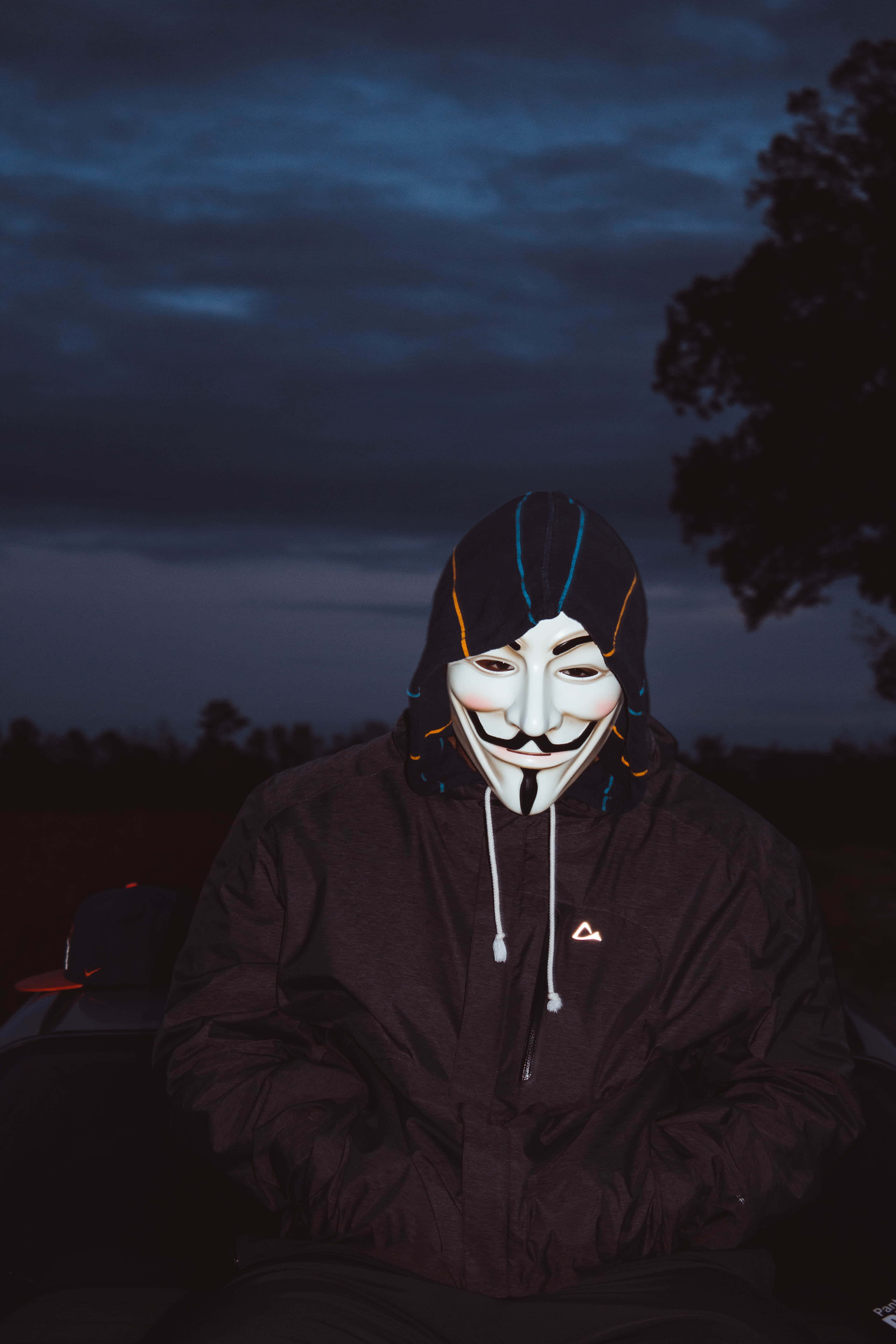 戴著 Vendetta 面具的男子站在陰鬱天空下 壁紙 — 一個人戴著白色蓋伊·福克斯面具和深色連帽衫，坐在陰沉的暮色天空和剪影樹木前。