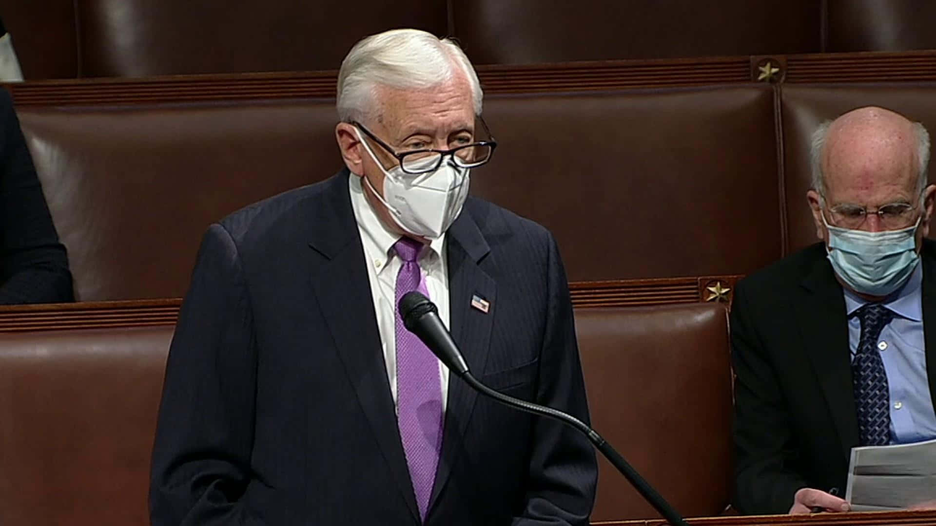 戴著口罩的斯泰尼·霍耶 壁紙 — 一位年長男性身穿深色西裝和紫色領帶，在正式的立法會議廳內戴著白色口罩對著麥克風發言。