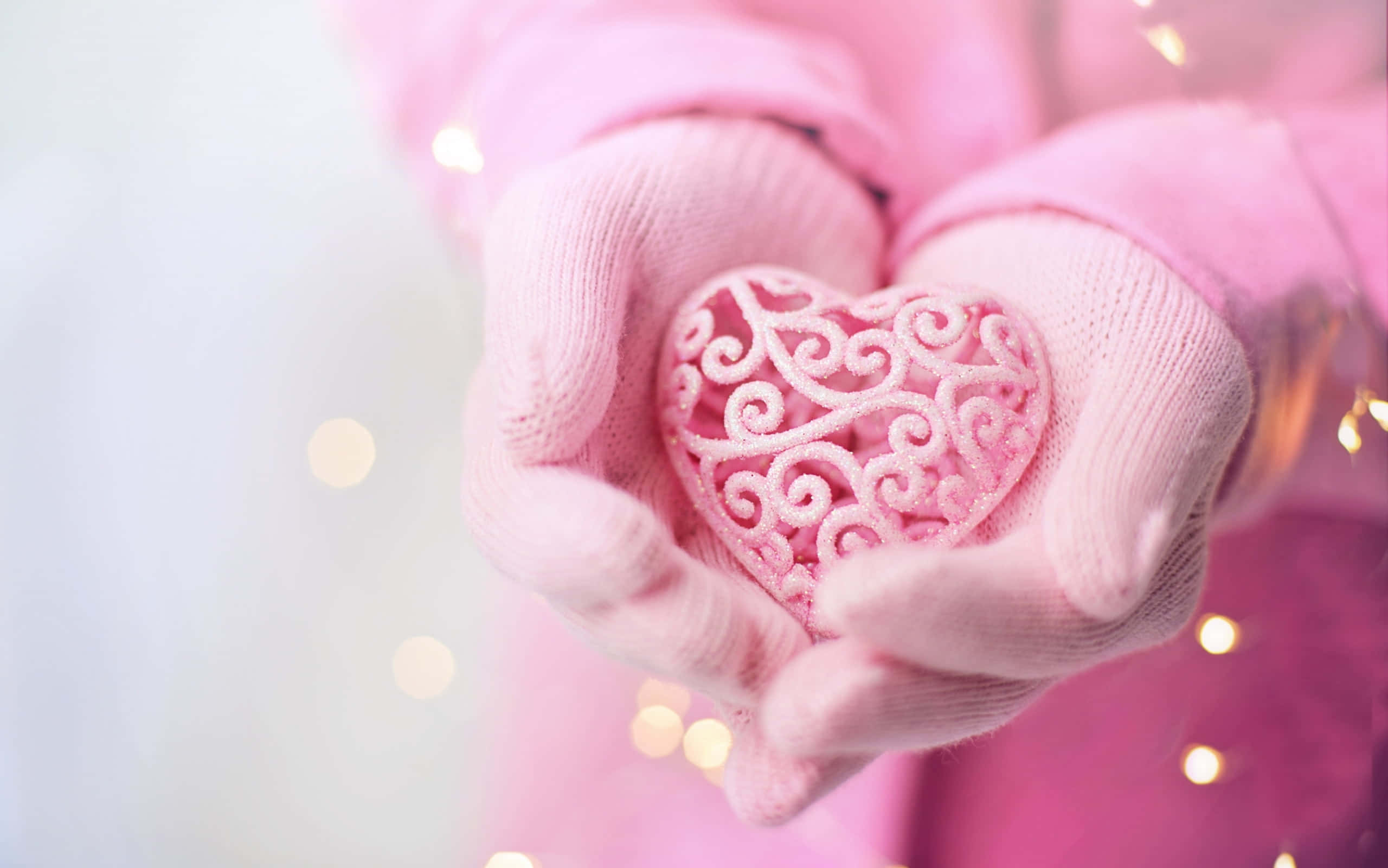 Mittens 桌布 壁紙 — 戴著手套的雙手，在溫暖燈光的柔和散景中，輕捧著一個精緻的粉紅色鏤空愛心。