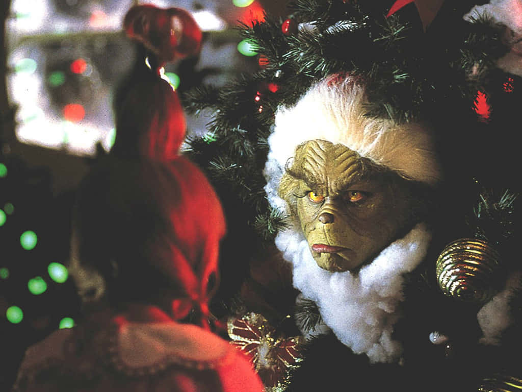 The Grinch 桌布 壁紙 — 戴著聖誕帽的鬼靈精從裝飾好的聖誕樹後面憤怒地窺視，一個穿紅衣的孩子在看著。