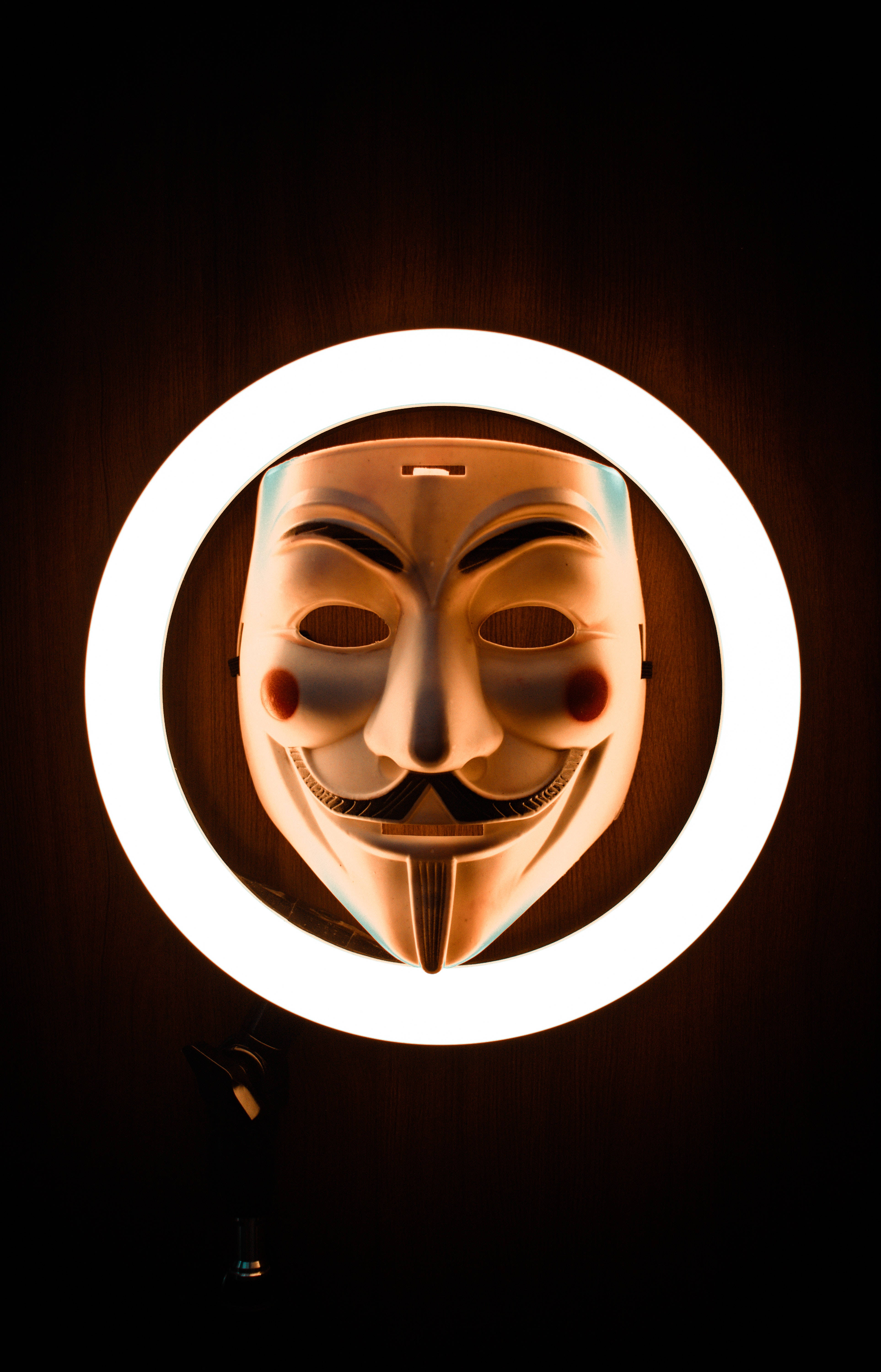 戴著面具的神秘黑客，環形燈照亮 壁紙 — 一個白色嘅 Guy Fawkes 面具，被明亮嘅環形燈照亮，背景係深色木牆。