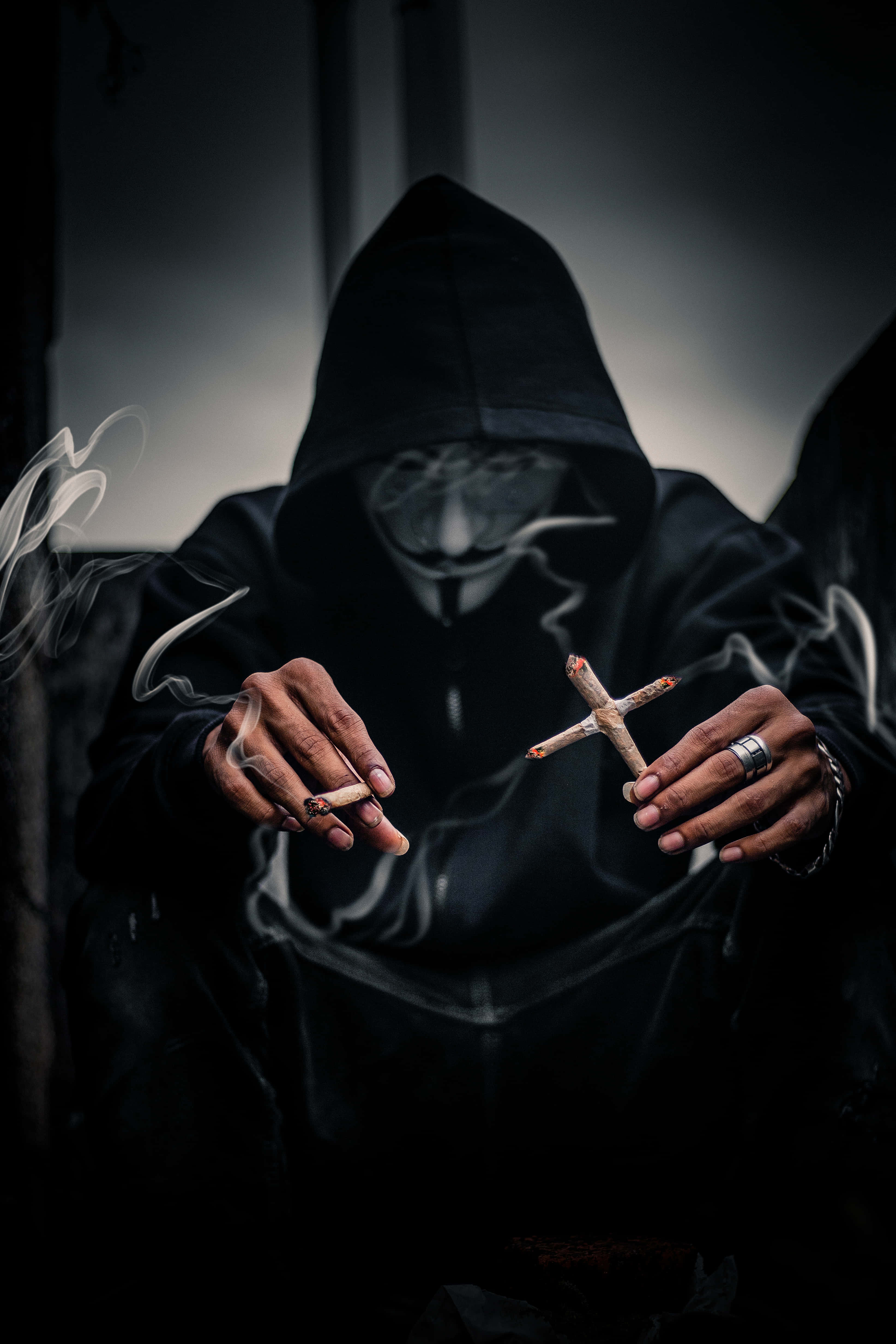 戴蓋伊·法威克面具的男子手持十字煙草 壁紙 — 一個戴著 Guy Fawkes 面具的兜帽人物在昏暗燈光下抽著十字形大麻煙，同時手持一根香煙。