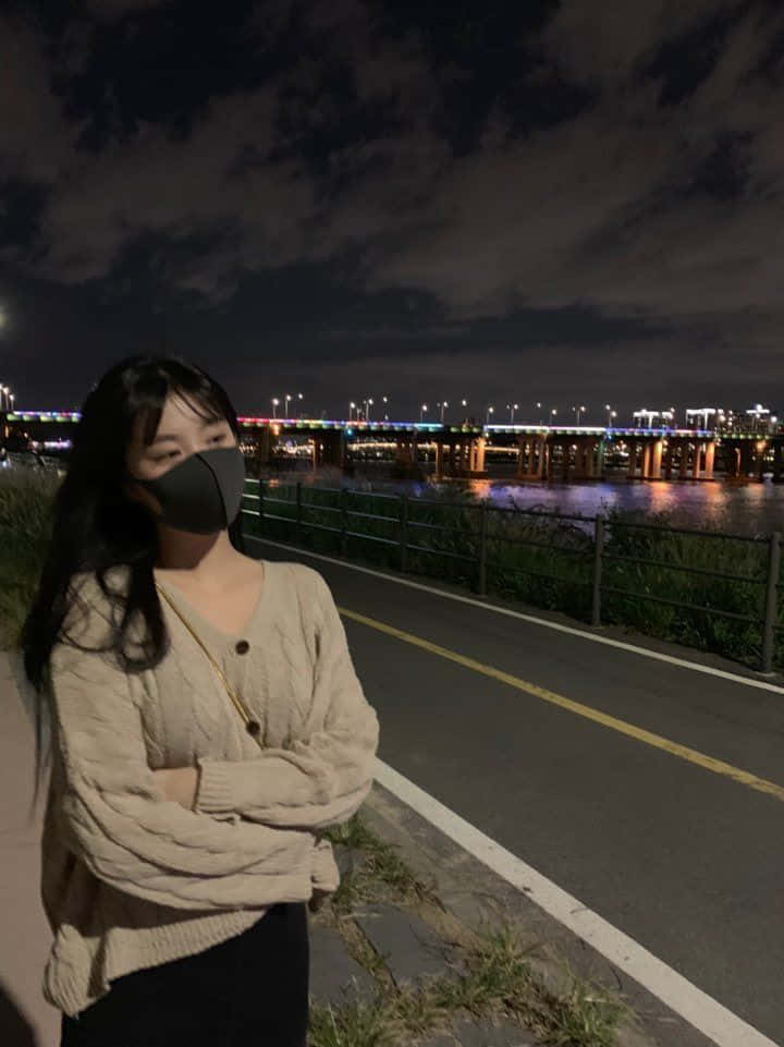 晚上，戴著口罩的人站在燈光璀璨的橋下的河邊。