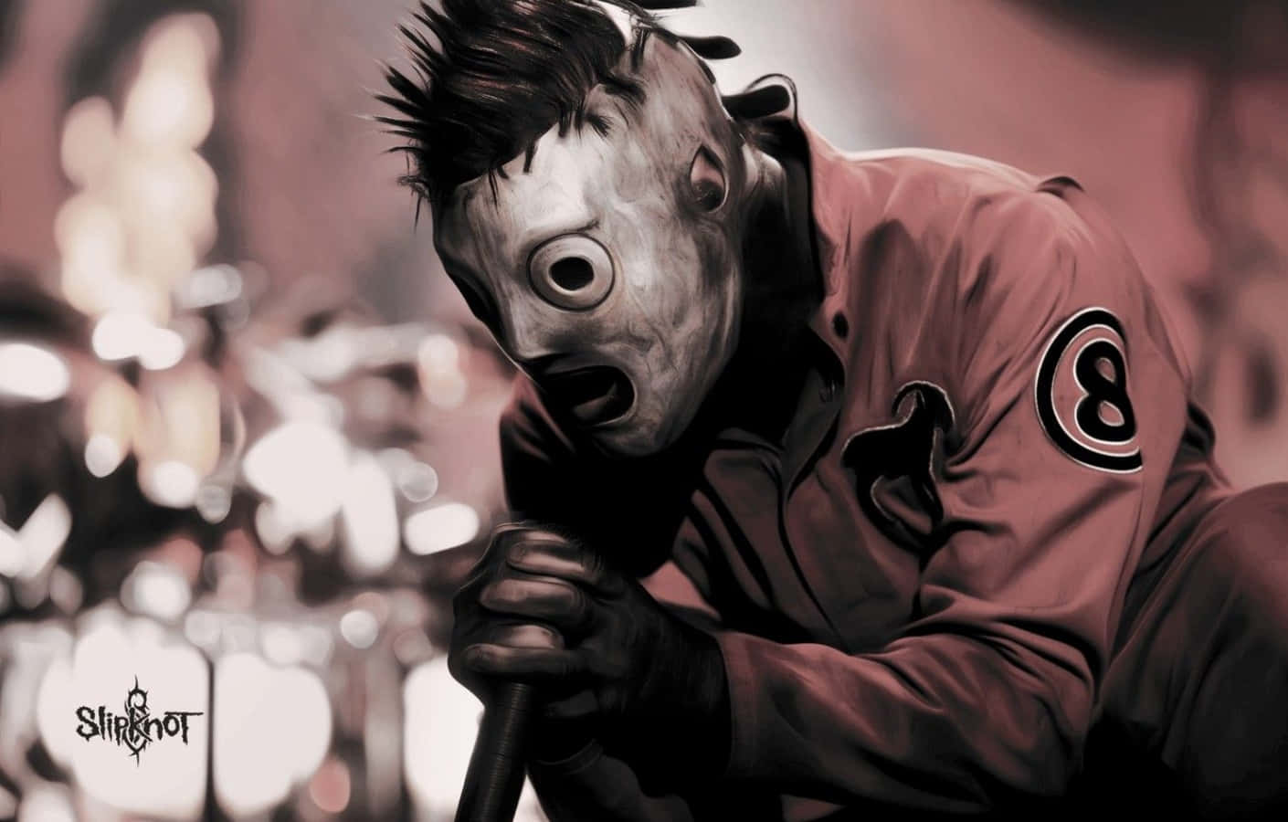 戴面具的Slipknot樂隊演唱藝術家桌面圖像 壁紙 — 一個戴著面具、身穿紅色外套的人物，在模糊的舞台背景下，熱情地對著麥克風唱歌。