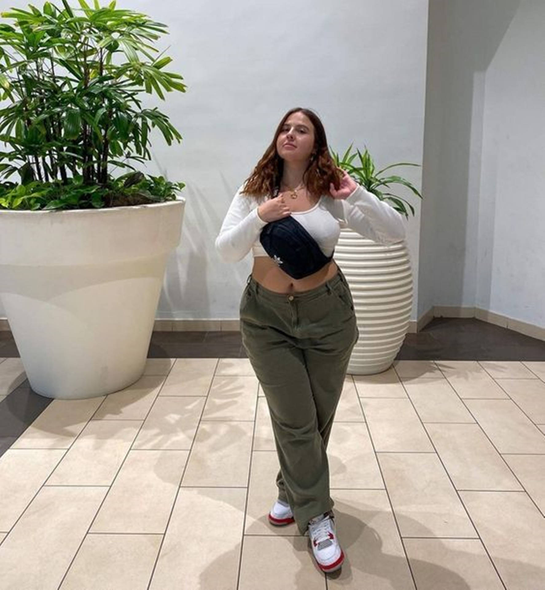年輕女性身穿橄欖色長褲和白色上衣，在室內盆栽植物附近佩戴腰包。