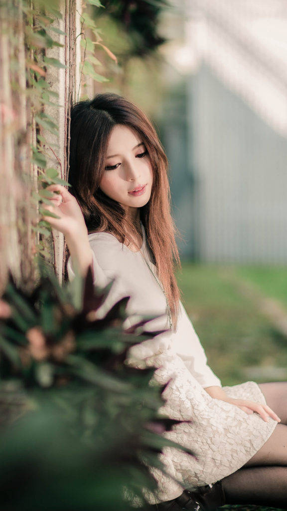 戶外時尚少女 壁紙 — 一位年輕女子身穿奶油色蕾絲連衣裙，在柔和自然光線下靠在一面爬滿藤蔓的牆上。