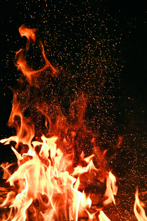 戶外篝火火焰背景 壁紙 — 鮮豔的橙色和黃色火焰在漆黑的背景上猛烈上升，空氣中漂浮著發光的餘燼。