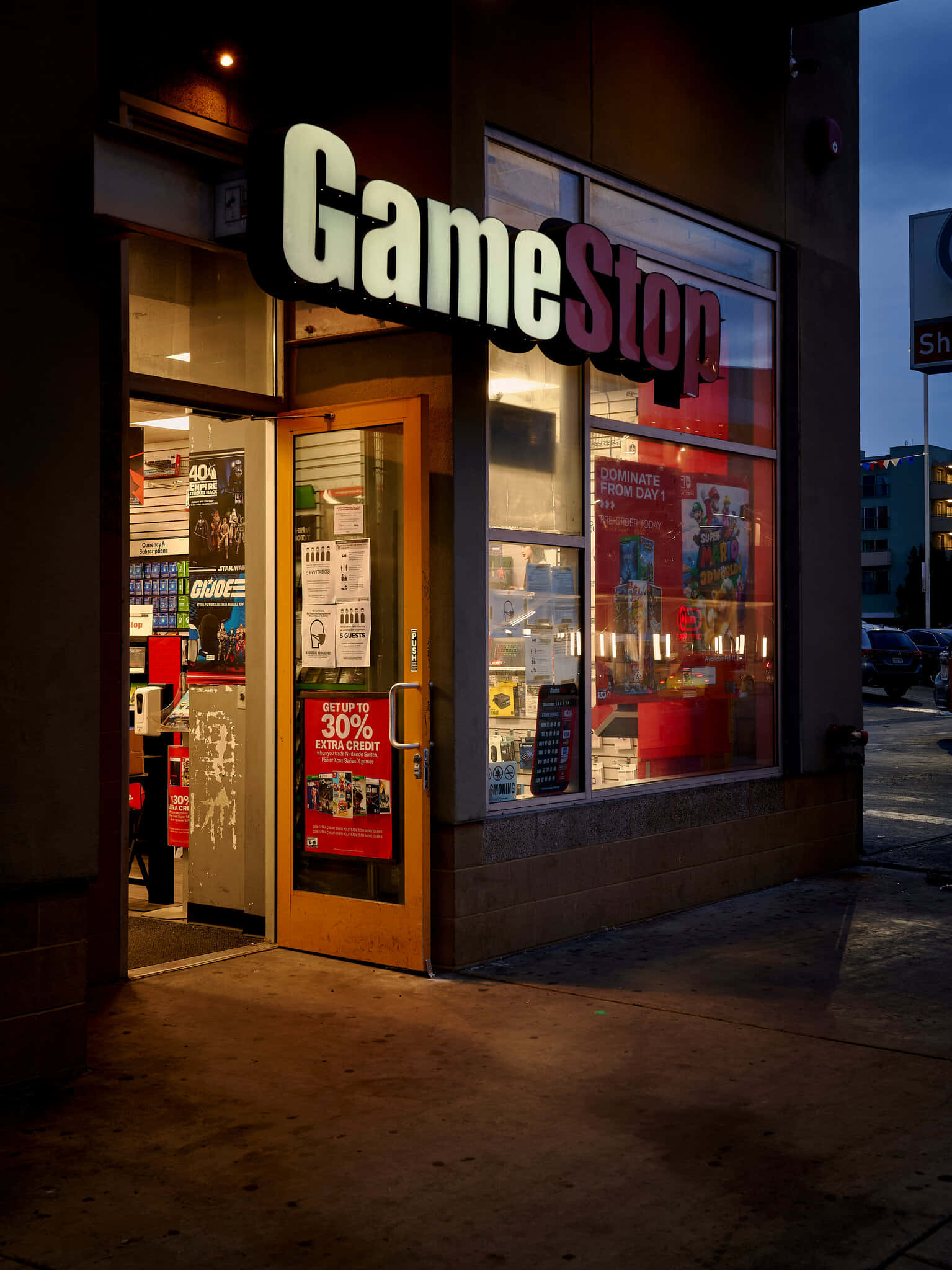 所有您最愛的遊戲都在一個地方 壁紙 — 黃昏時分的GameStop商店入口，溫暖的室內燈光透過玻璃窗和木門閃爍。