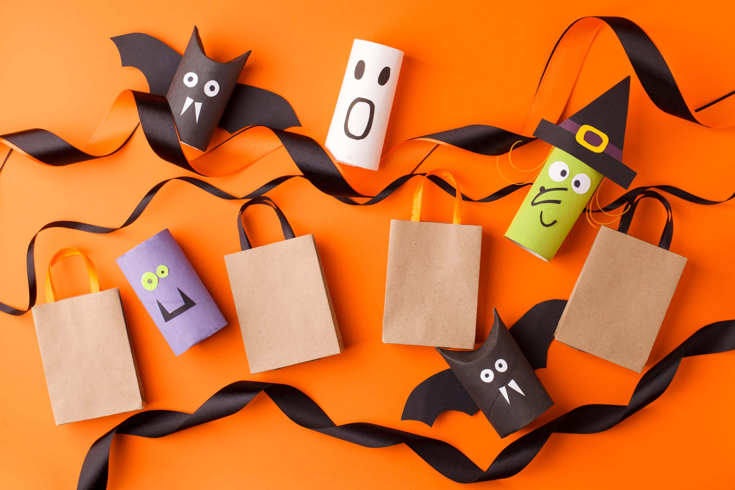 Halloween Crafts 桌布 壁紙 — 手作萬聖節小食，裝在紙袋中，紙袋上有蝙蝠、鬼魂、女巫臉孔，背景為亮橙色，配有黑色絲帶。