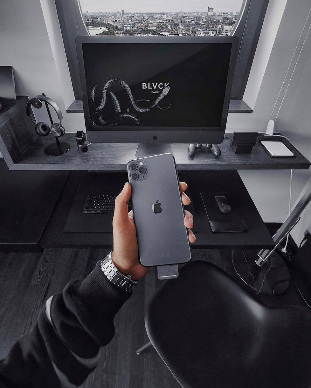 Blvck Paris iPhone 11 Pro Max 壁紙 — 手在時尚黑色辦公桌前拿著深灰色iPhone，顯示器顯示蛇標誌和透過窗戶的城市景觀。