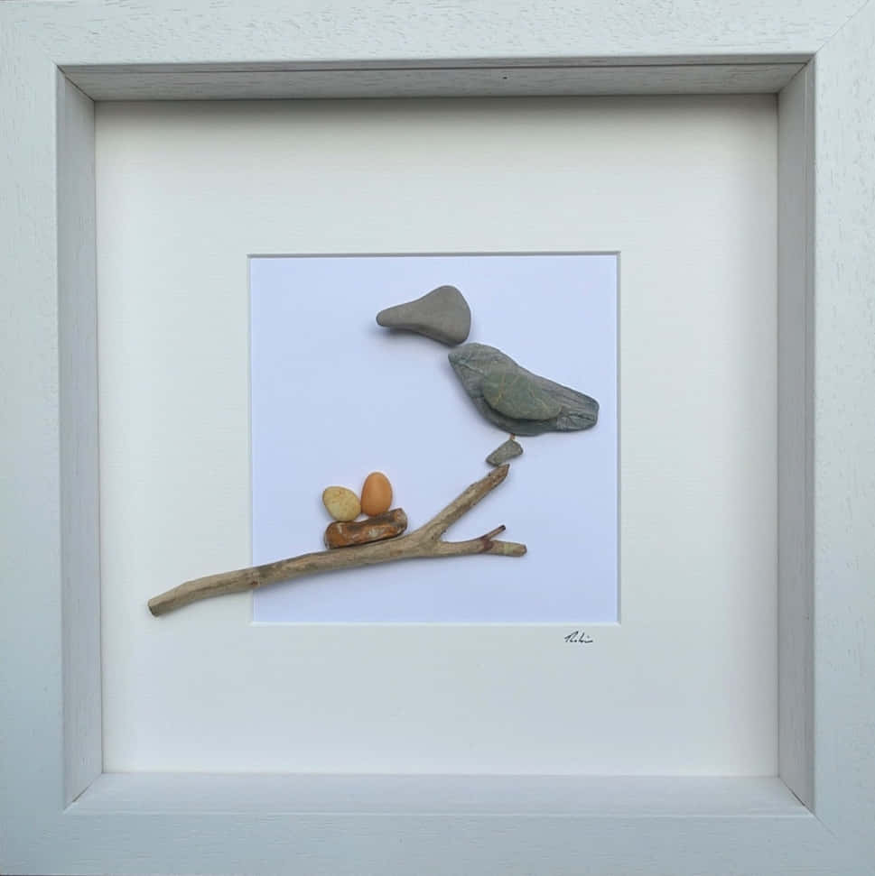 手工 Pebble Art 照片，搭配貝殼與心形設計 壁紙 — 一幅用光滑石頭和浮木製成的裱框藝術品，描繪了一隻棲息在樹枝上的鳥，下方有兩個鳥蛋。