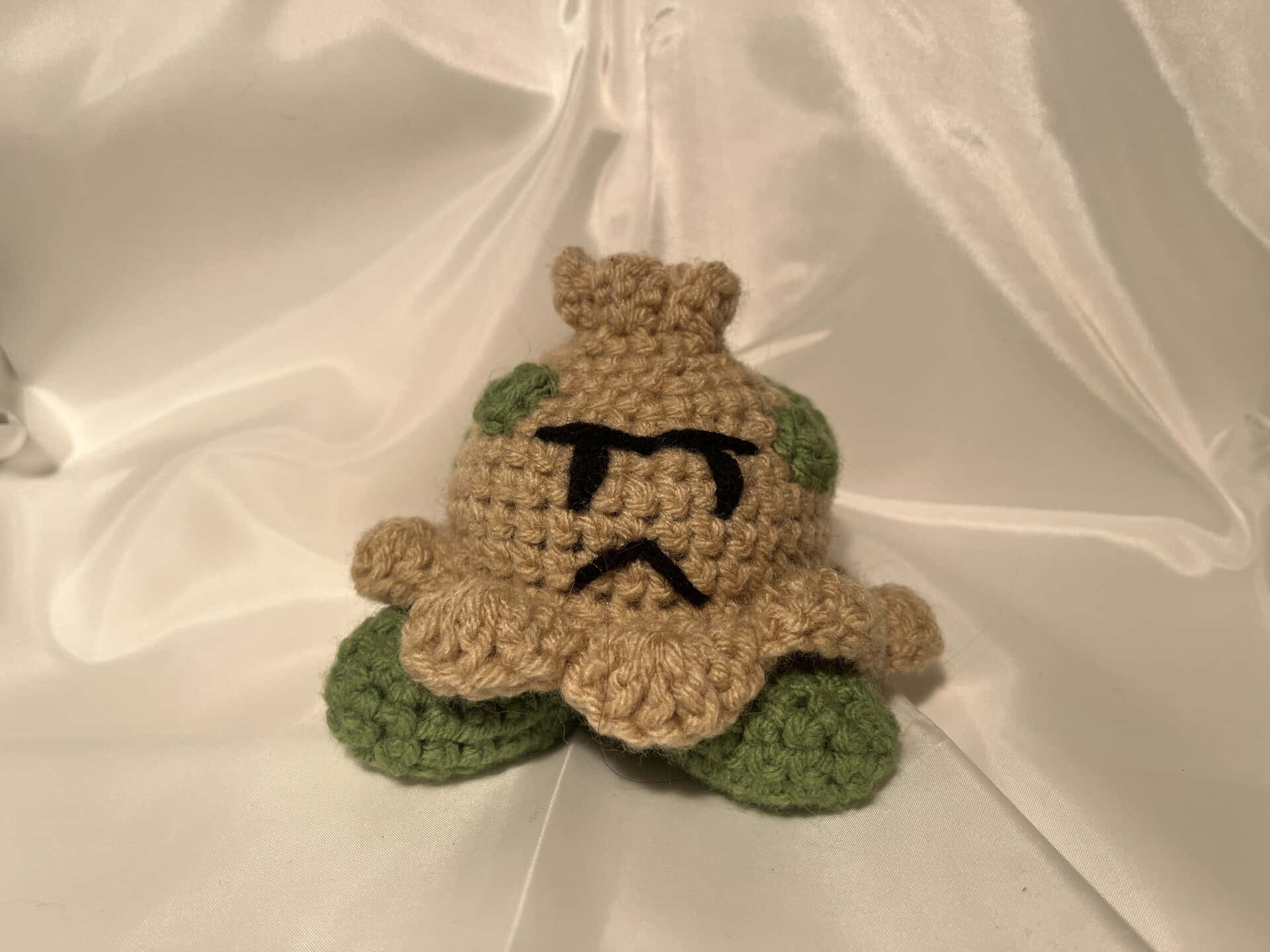 鉤織 Bulbasaur 毛公仔，帶有綠色葉子和黑色縫製的臉，放在白色緞面上。