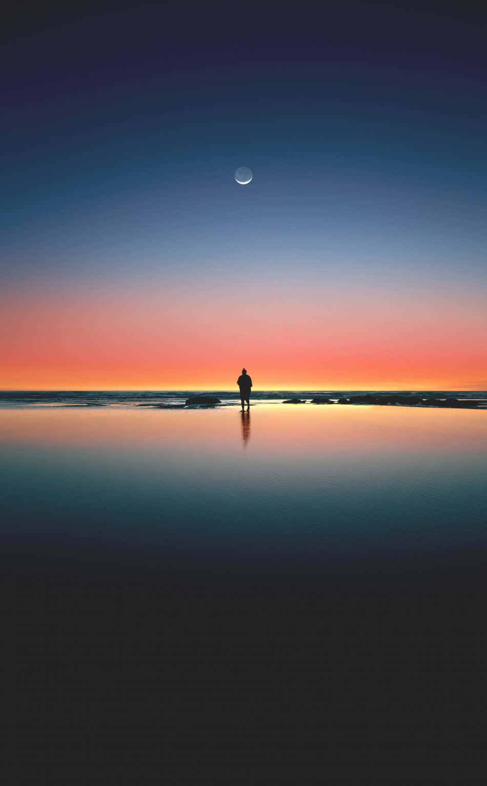 手持 iPhone，享受溫暖的日落海灘時光 壁紙 — 黃昏時分，一個孤獨的身影站在濕漉漉的海灘上，一彎新月掛在漸變色的天空中，倒映在平靜的水面上。