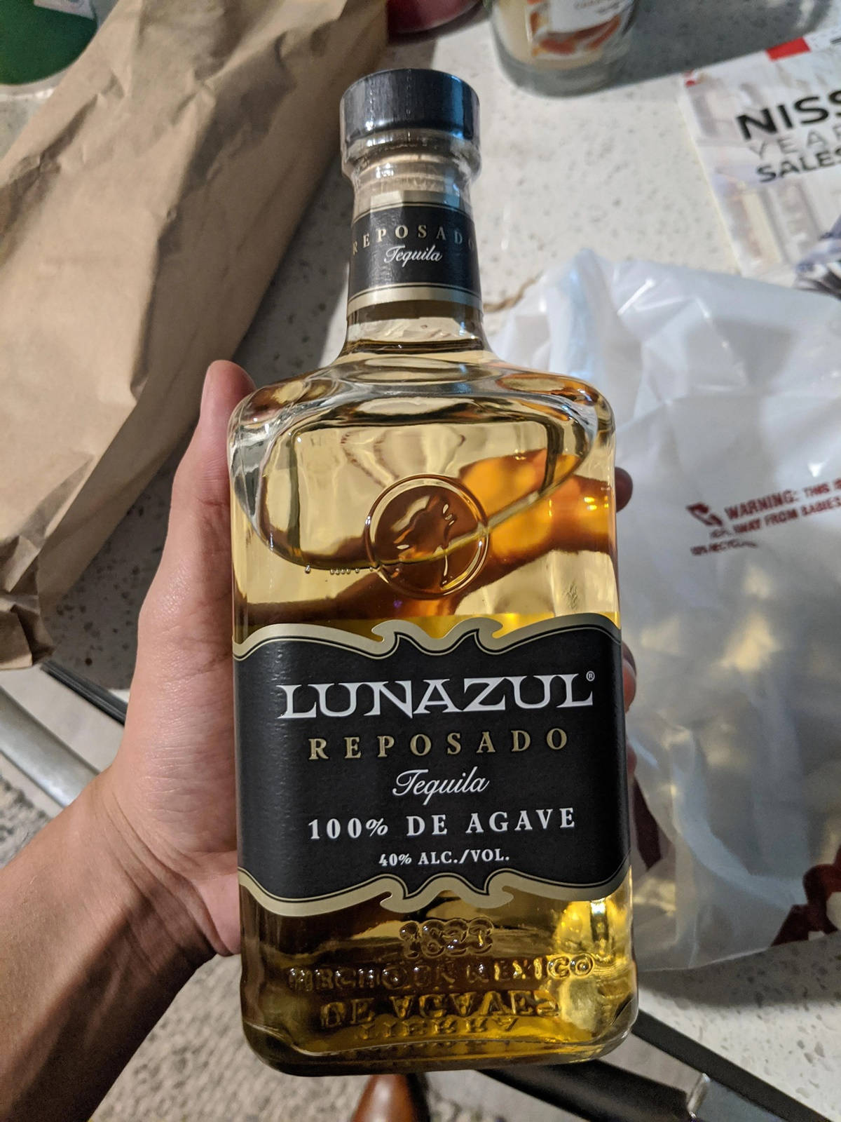 手持Lunazul Reposado龍舌蘭酒 壁紙 — 一隻手拿著一瓶Lunazul Reposado龍舌蘭酒，金色液體透過透明玻璃清晰可見，背景是雜亂的檯面。