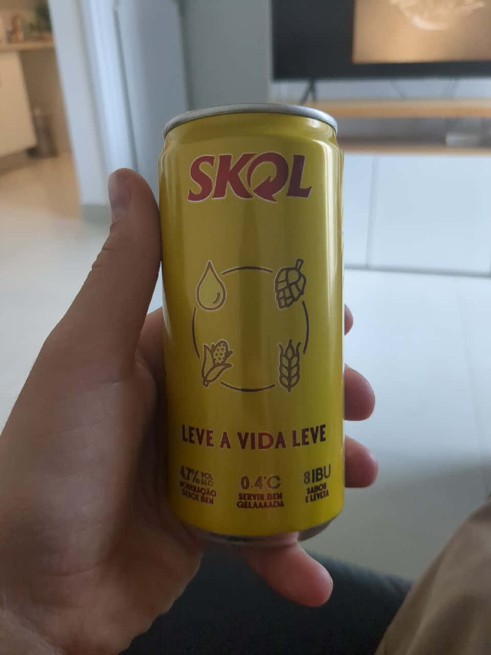 手持Skol啤酒罐 壁紙 — 一隻手拿著一個亮黃色的Skol啤酒罐，上面有啤酒花和小麥圖標，背景是模糊的室內環境。