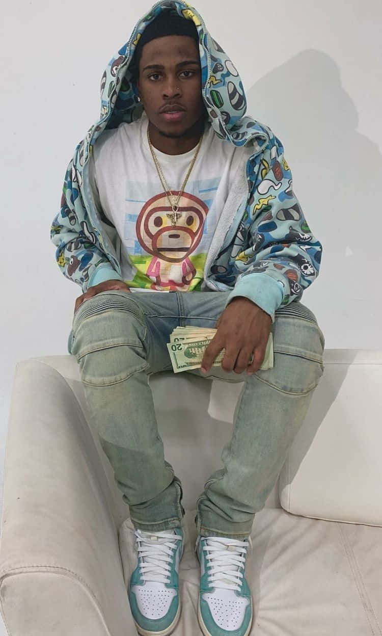 手持大量現金的 dope boy 壁紙 — 一名身穿彩色連帽衫和牛仔褲的年輕男子坐在白色沙發上，手持一疊現金，穿著猴子圖案T恤和青綠色運動鞋。