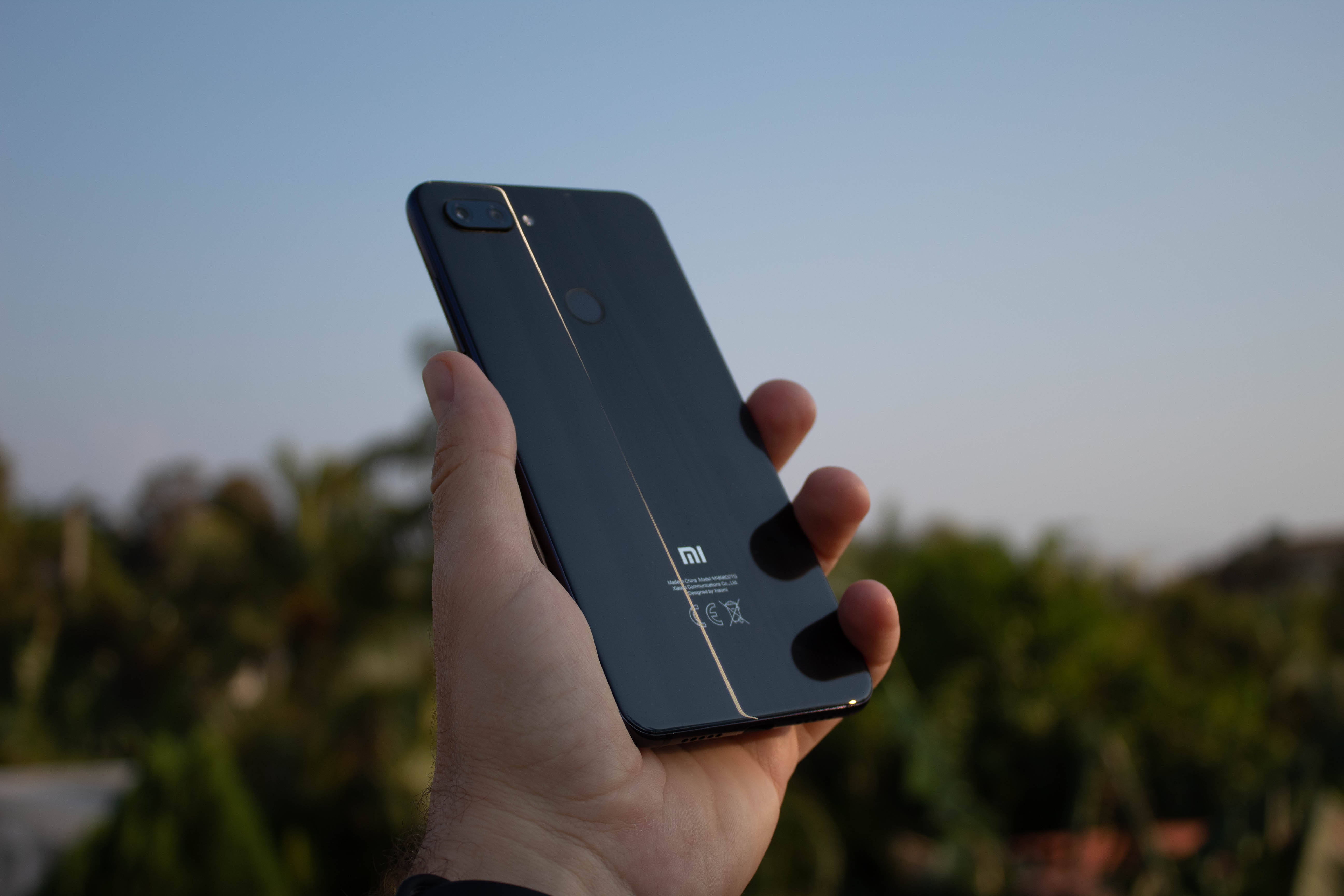 手持小米智能手機 壁紙 — 一隻手拿著一部時尚的黑色Xiaomi智能手機，背景是柔和的戶外環境，帶有溫暖的傍晚光線。