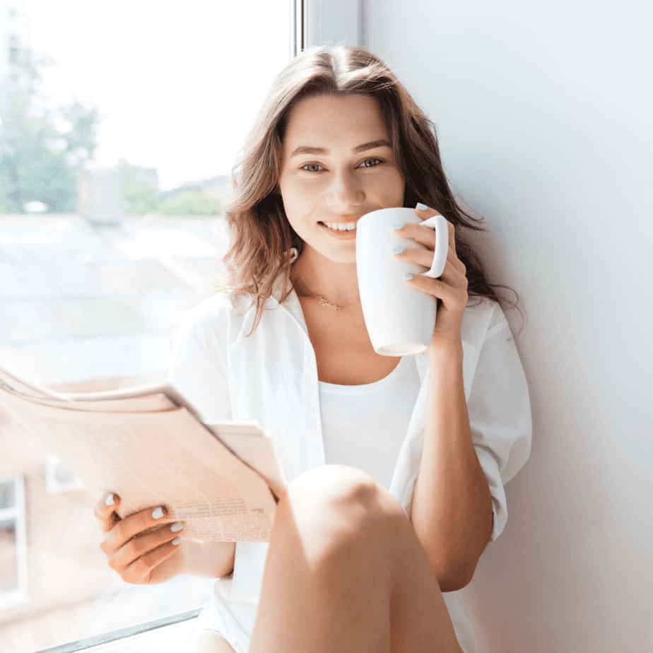 手持杯子的快樂女子畫面 壁紙 — 一位穿著白色衣服的微笑女士靠在窗邊，一邊喝咖啡一邊看報紙。