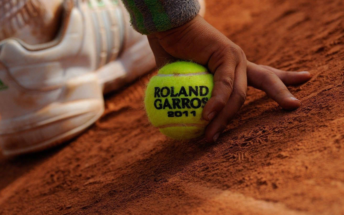 手持法國公開賽網球球的人 壁紙 — 一隻手抓住一個鮮黃色的Roland Garros 2011網球，在紋理紅土球場上，旁邊有一隻白色運動鞋。