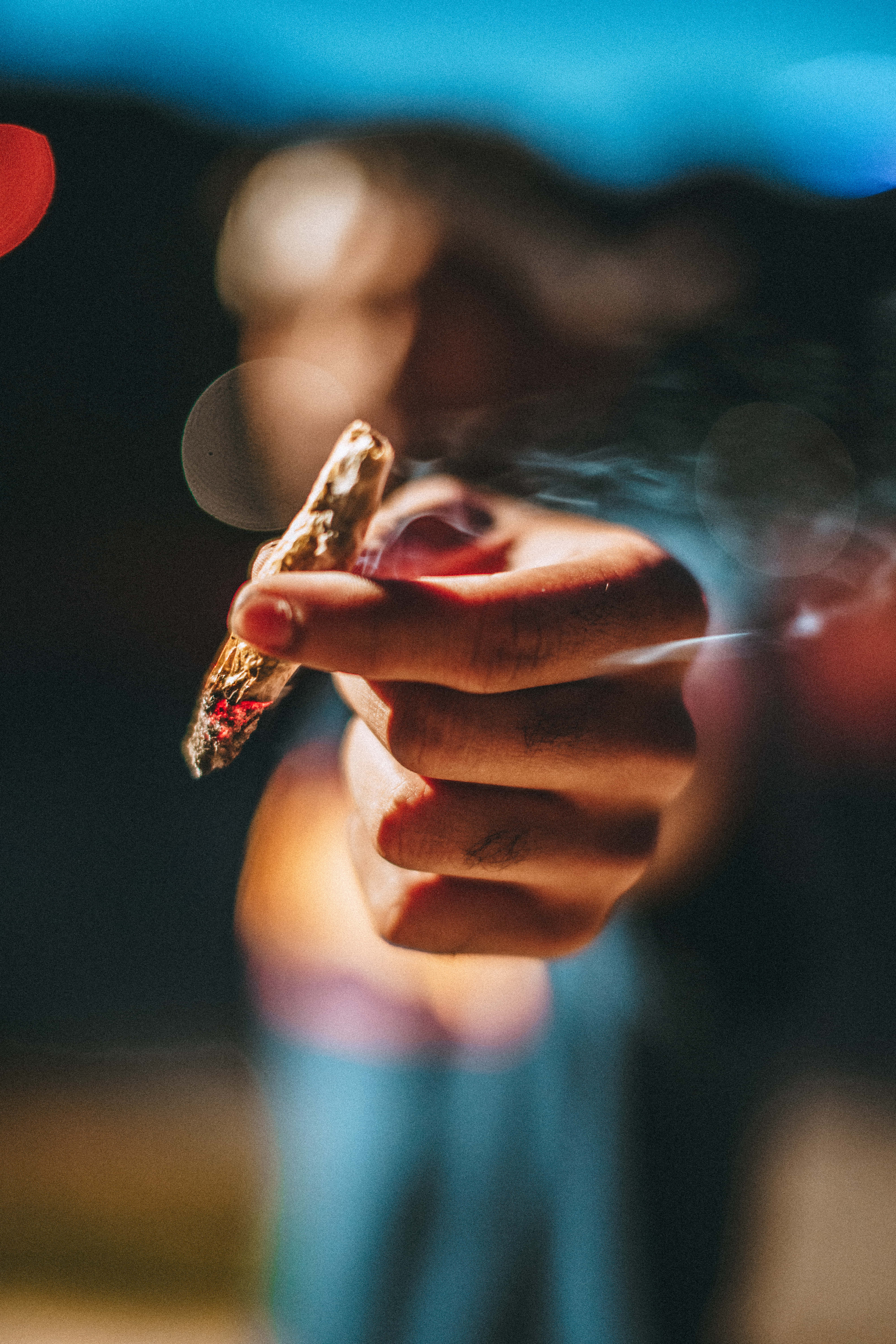手持煙草的特寫 壁紙 — 一隻手喺模糊背景、散景燈光同上升煙霧中揸住一支點燃嘅大麻煙。