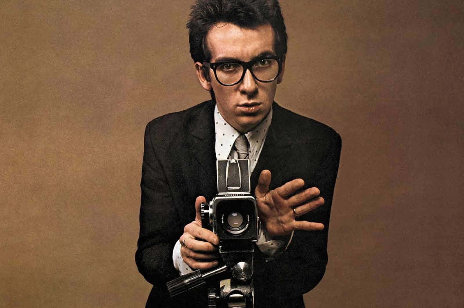 手持老式相機的年輕Elvis Costello 壁紙 — 一名身穿深色西裝、戴眼鏡的男子，在柔和的棕色背景下，以專注的眼神手持一部復古相機。