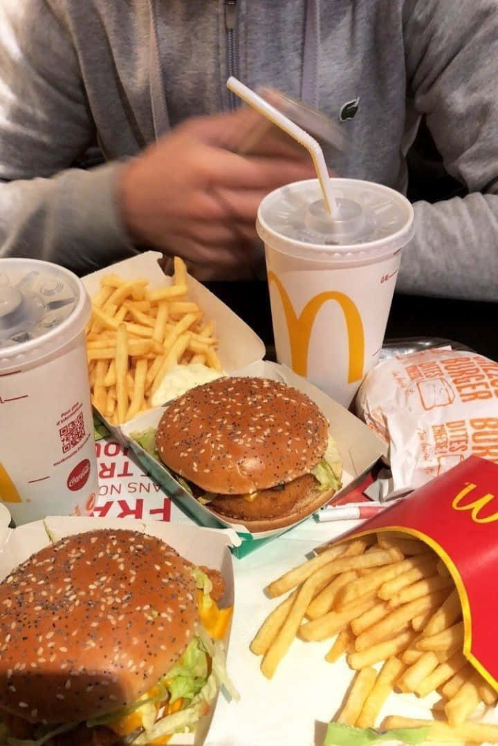 手持麥當勞餐點的男子 壁紙 — 麥當勞餐點的近距離拍攝，桌上放有兩個漢堡、薯條和飲品，背景有一名穿著灰色連帽衫的人。