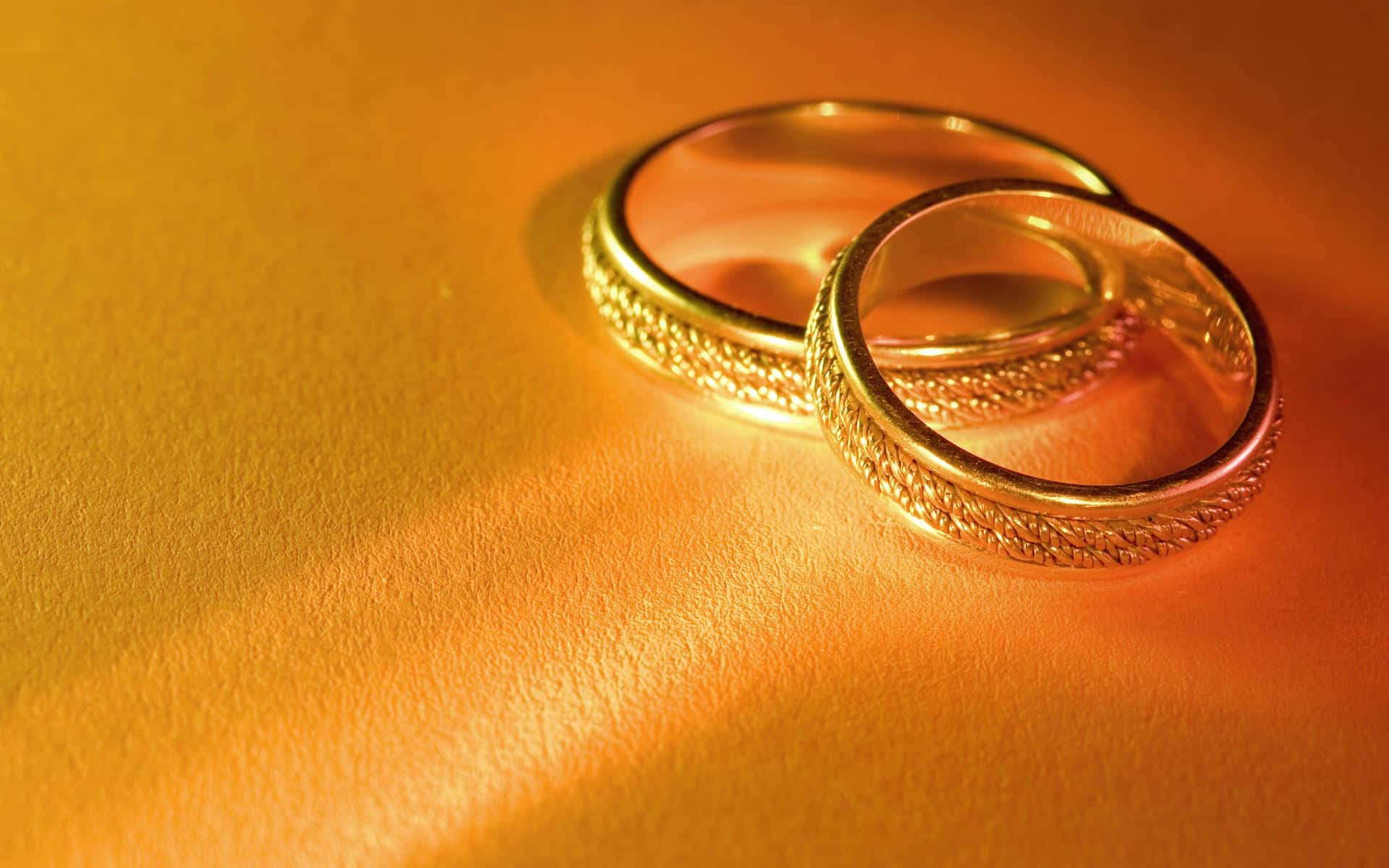 手指上閃耀的銀色戒指 壁紙 — 兩隻帶有紋理的金色結婚戒指，放置在溫暖的金色表面上，沐浴在柔和的方向光中。