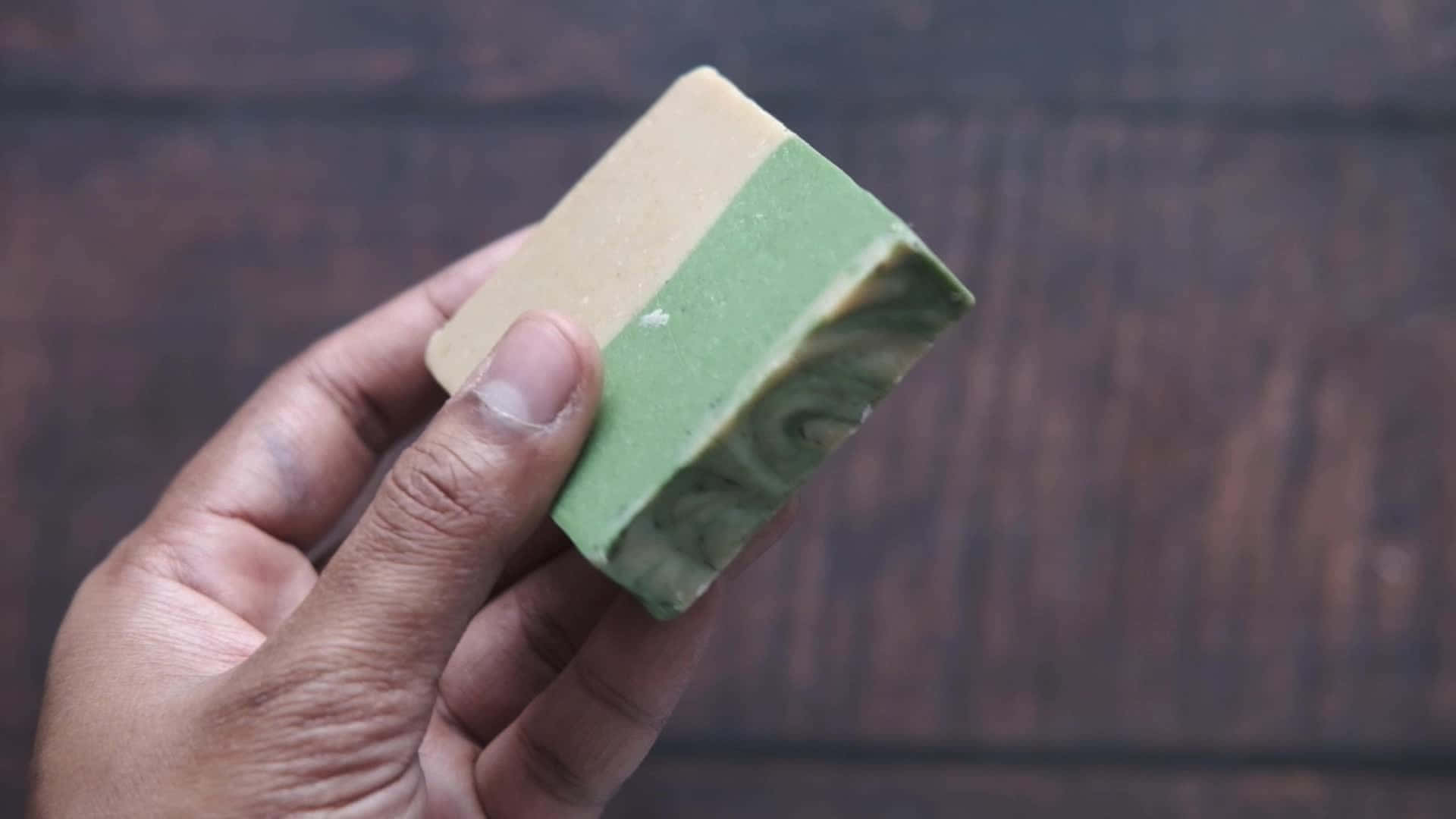 手握 Lush 香皂 壁紙 — 一隻手拿著一塊帶有綠色漩渦的雙色肥皂，背景是深色木材。