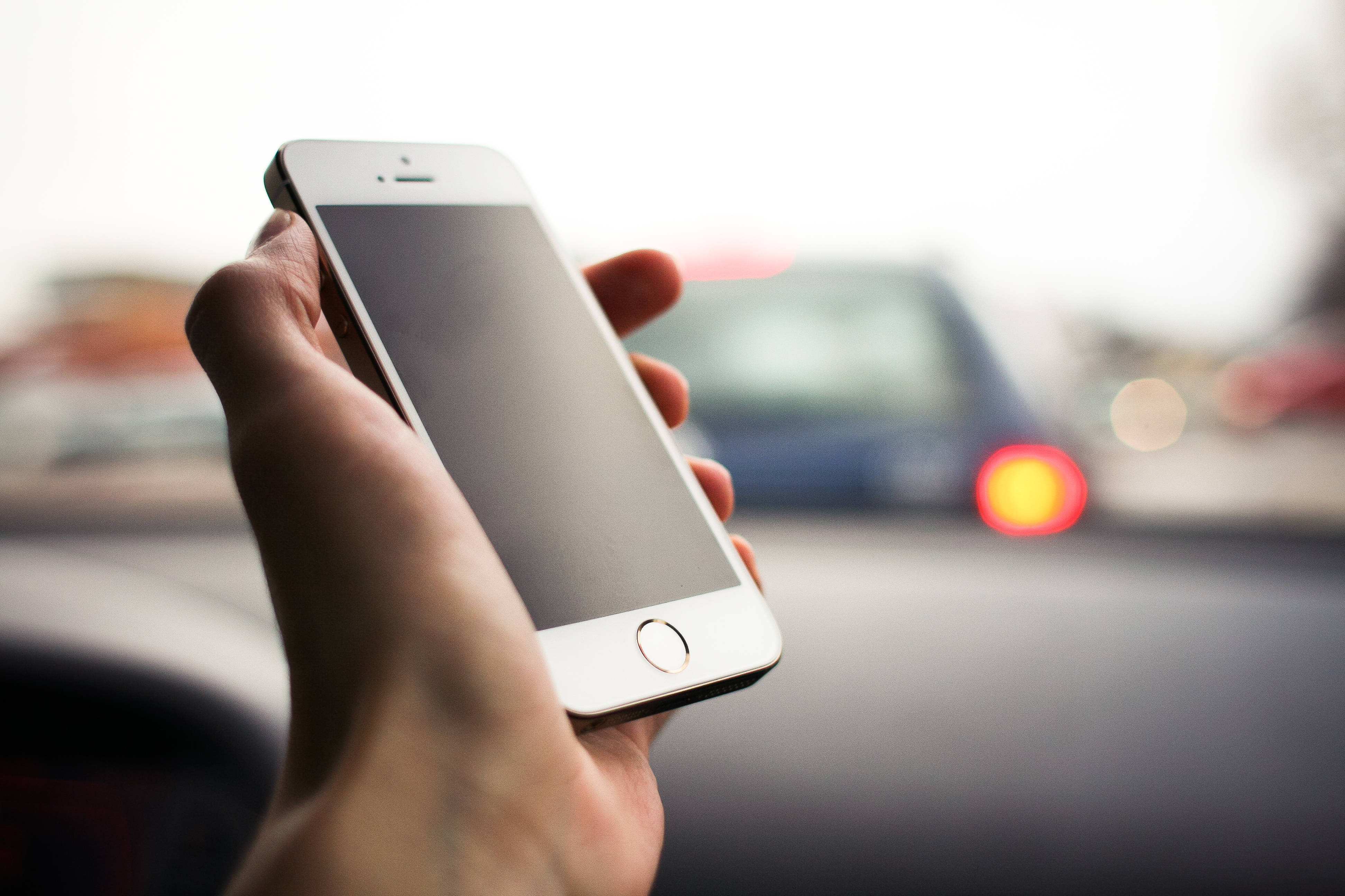 手握原版 iPhone 5 壁紙 — 一隻手在車內拿著一部白色智能手機，背景模糊，顯示紅色尾燈和經過的交通。