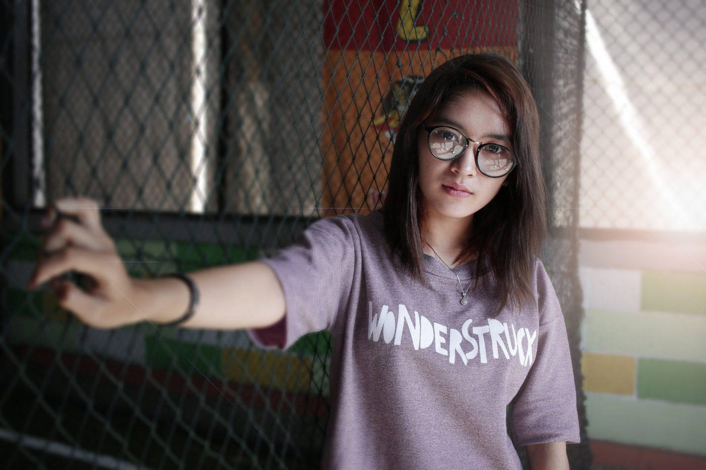 手搭在柵欄上的態度女孩 壁紙 — 一位戴眼鏡、身穿'WONDERSTRUCK' T恤的年輕女士在柔和陽光下靠著鐵絲網圍欄。