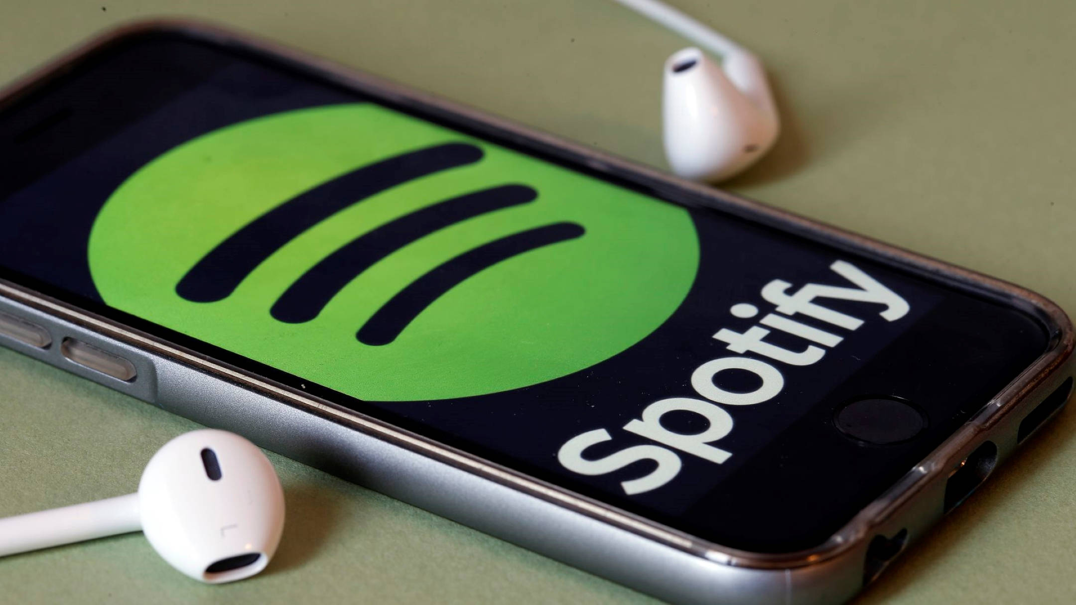 手機上的 Spotify 音樂 壁紙 — 一部智能手機顯示Spotify標誌，白色耳機放在旁邊的柔和綠色表面上。