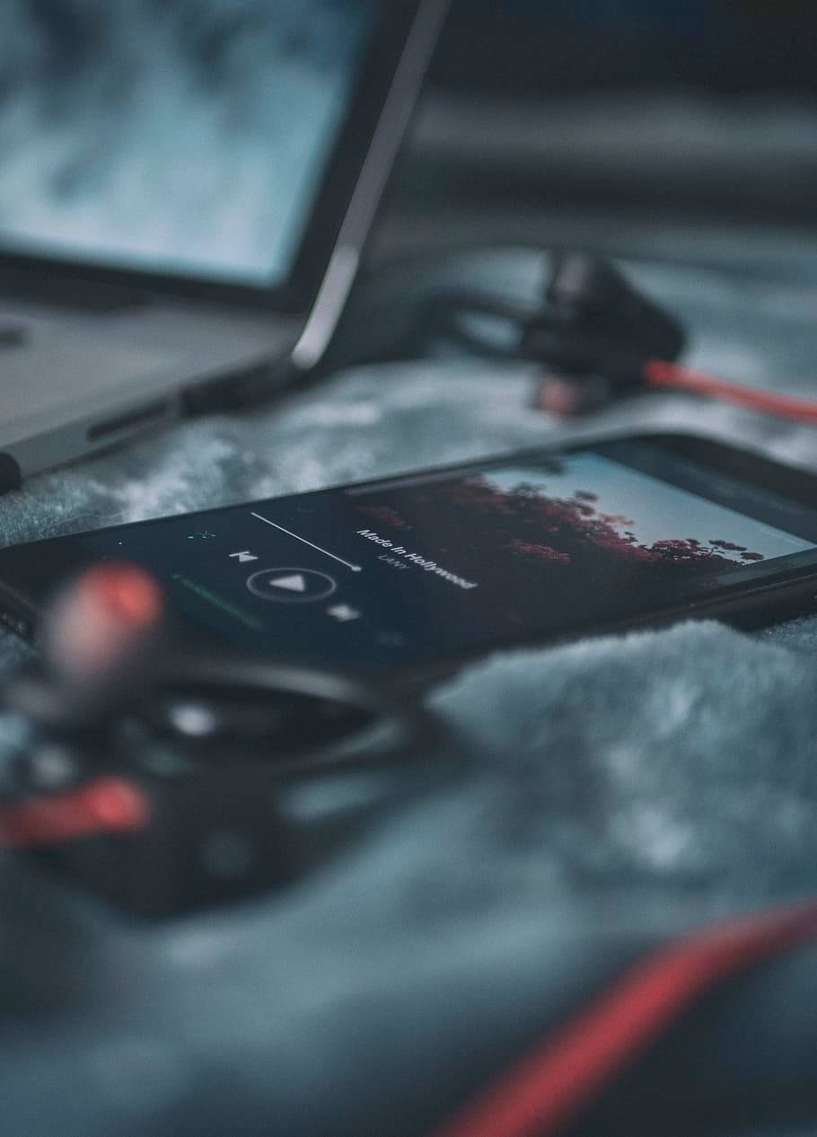 手機上的美學音樂體驗 壁紙 — 一部正在播放音樂的智能手機放在柔軟的灰色毛毯上，旁邊有一部手提電腦和紅色耳塞，營造出舒適的室內環境。