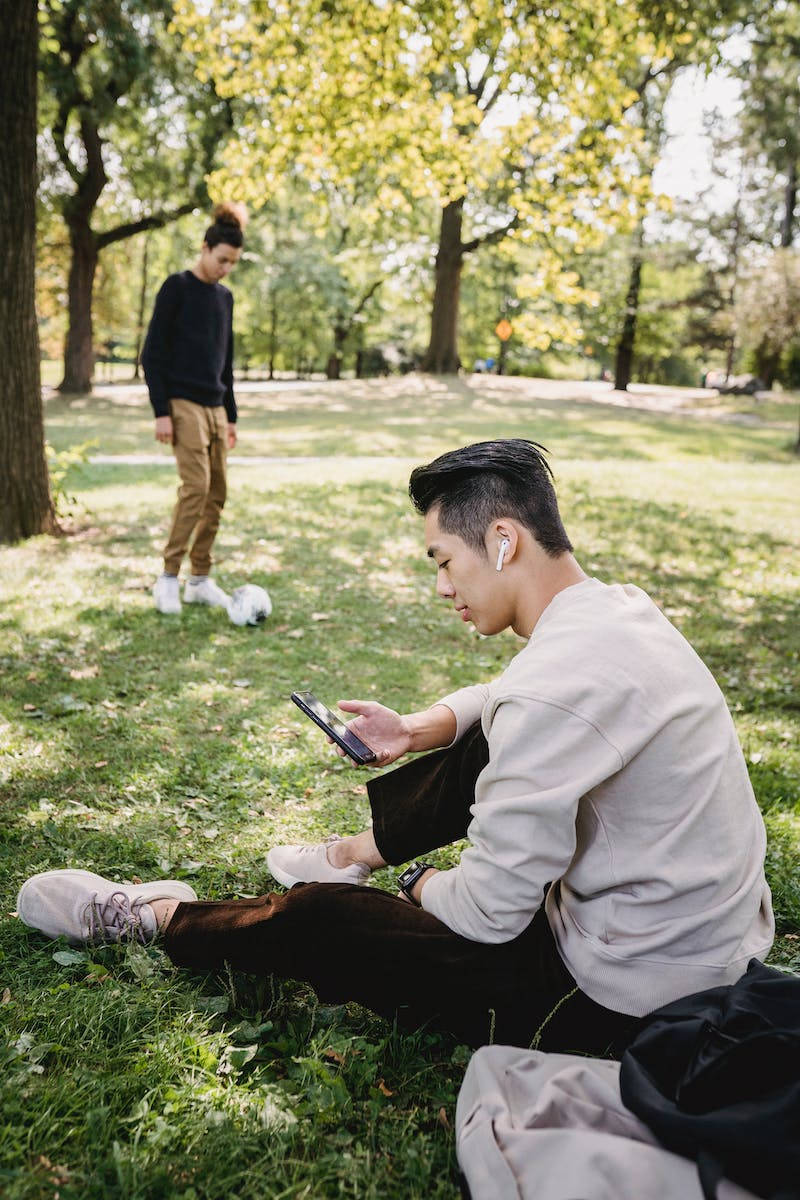 公園裡的兩個年輕男子，一個坐在草地上使用手機，另一個站著拿著足球