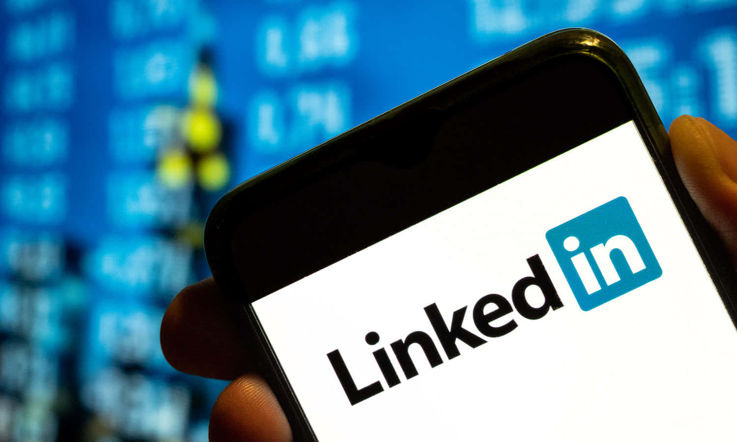 手拿著手機顯示LinkedIn應用程式，背後有股票市場數據。