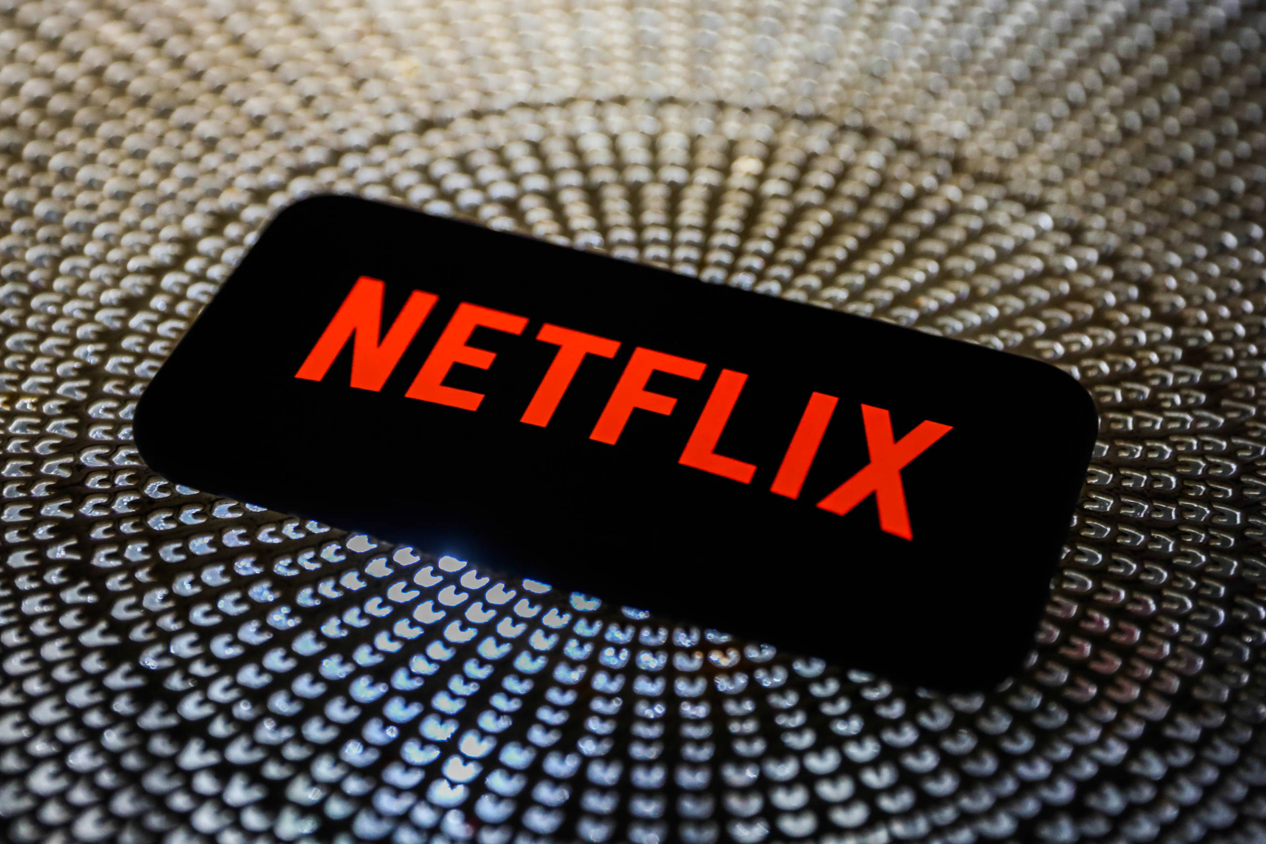 手機螢幕上的Netflix標誌 壁紙 — 一部顯示鮮紅色Netflix標誌的智能手機，放置在帶有放射狀圖案的紋理金屬表面上。