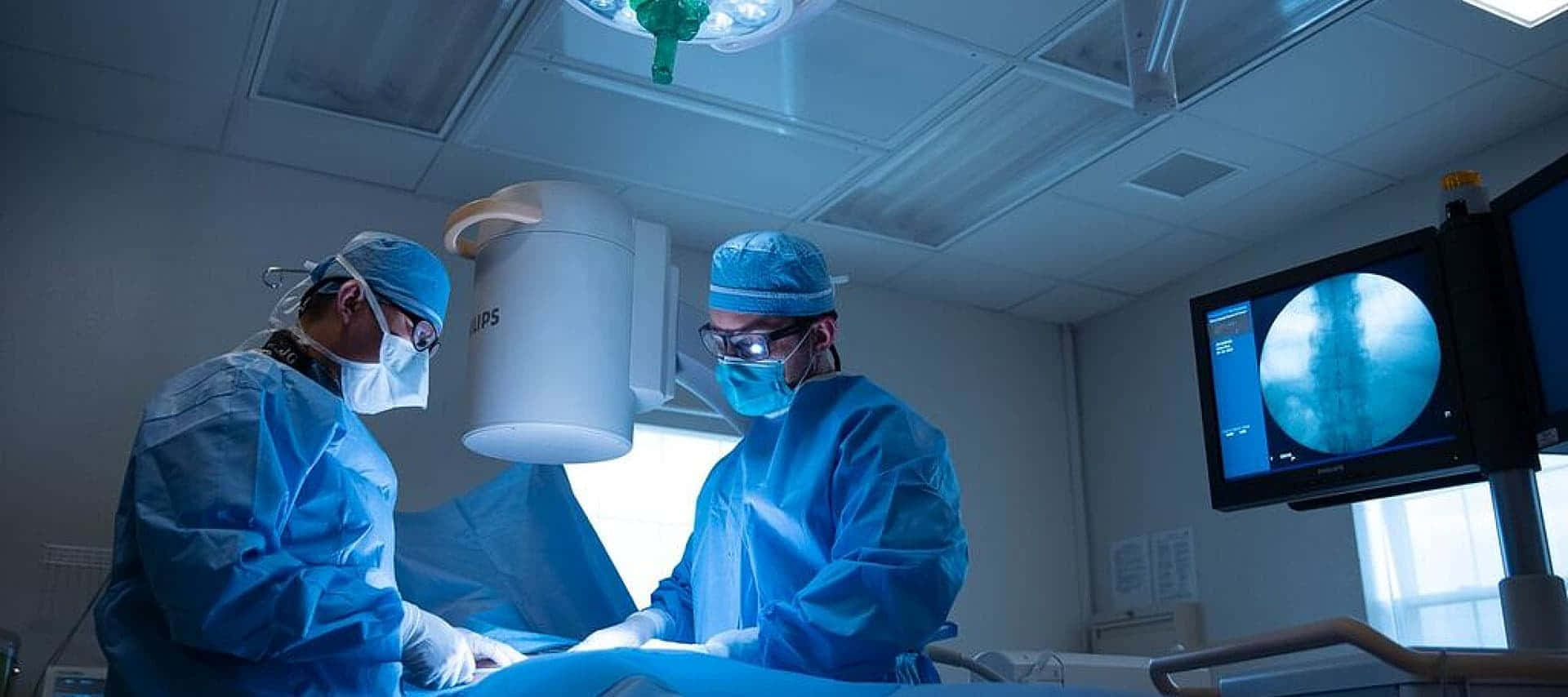 兩名身穿藍色手術衣的外科醫生在明亮燈光下進行手術。