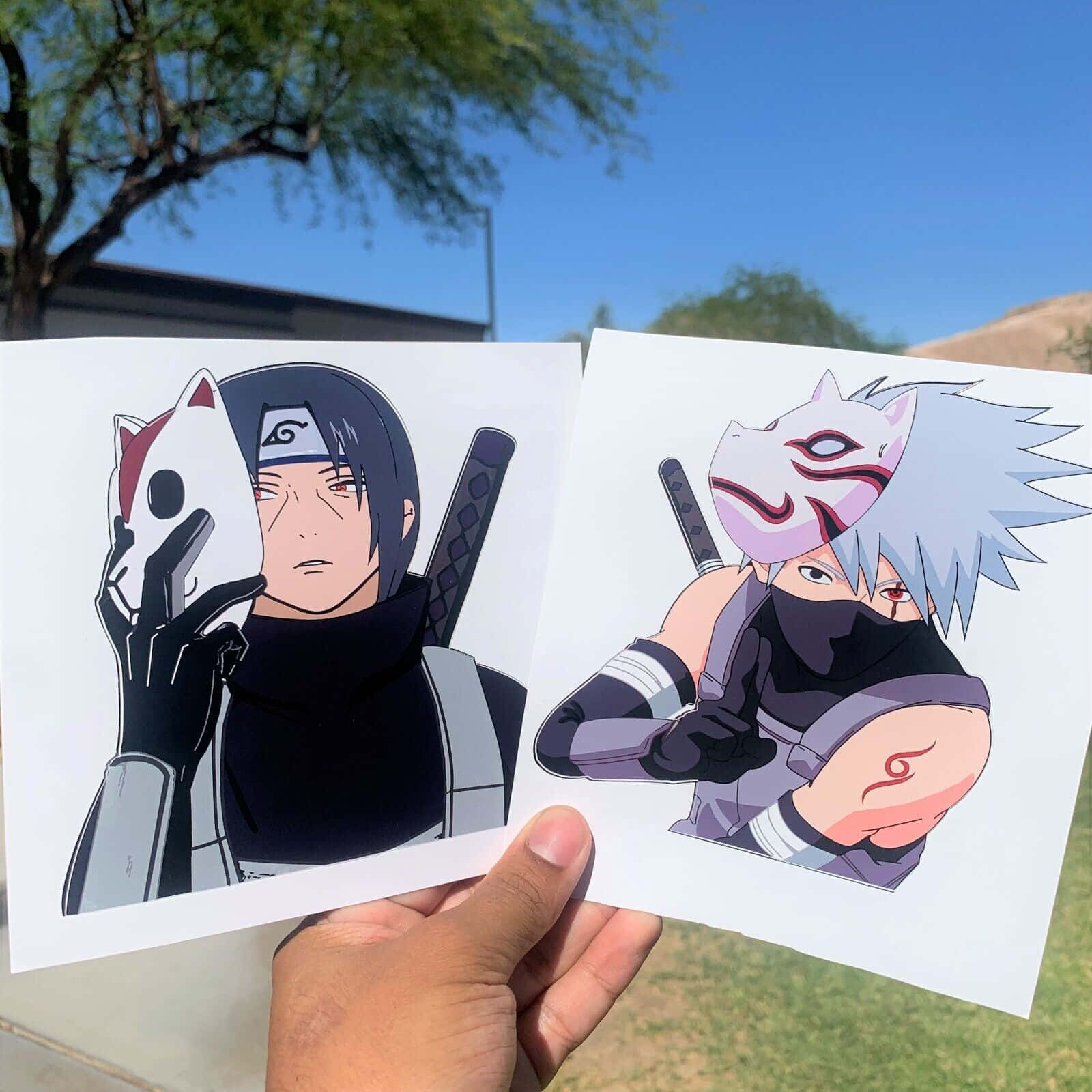 Kakashi And Itachi 桌布 壁紙 — 手裡拿著兩張戴面具的忍者角色動漫印刷品，背景是明亮的藍天和綠樹。