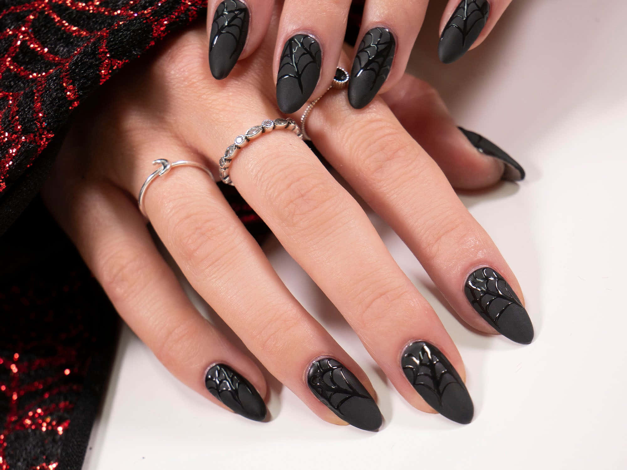 Halloween Nail Art 桌布 壁紙 — 手部特寫，帶有啞光黑色蜘蛛網指甲藝術和銀戒指，背景為紅色閃粉布料和白色表面。