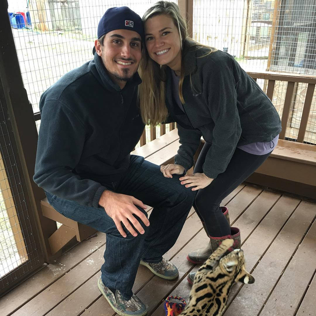 扎克·埃弗林與妻子在動物園 壁紙 — 一對微笑的情侶蹲在動物圍欄內的木甲板上，旁邊有一隻頑皮的豹貓。