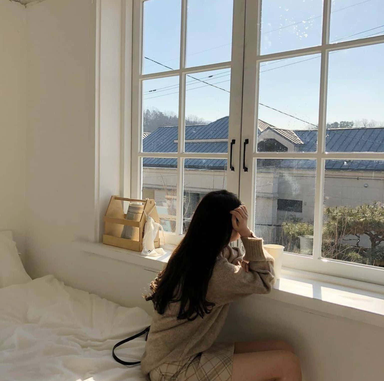打造寧靜綠洲的雅緻米色配色方案 壁紙 — 一位長黑髮的年輕女性坐在簡約臥室的明亮窗邊，凝視著晴朗藍天下屋頂的景色。
