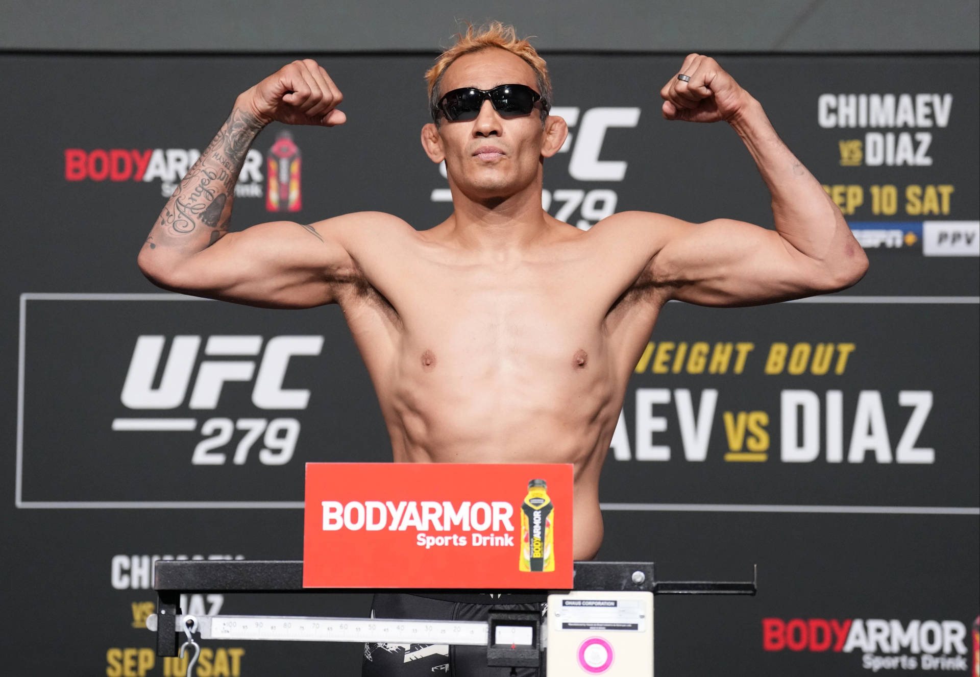 UFC拳手在稱重活動中赤裸上身彎曲肌肉，戴太陽眼鏡和紋身。