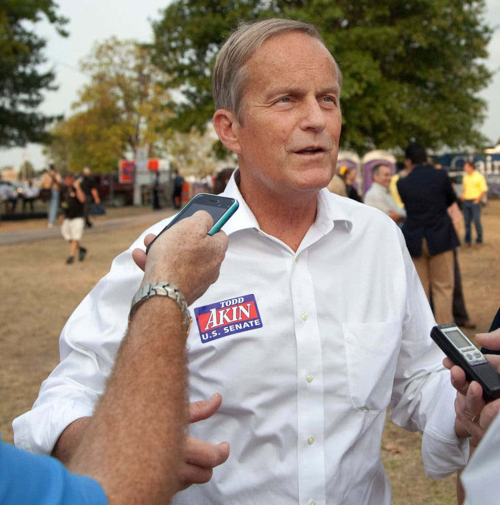 托德·阿金競選之路 壁紙 — 一名身穿白恤衫、佩戴 Todd Akin 美國參議院競選徽章的男子，在日間戶外向記者發言。