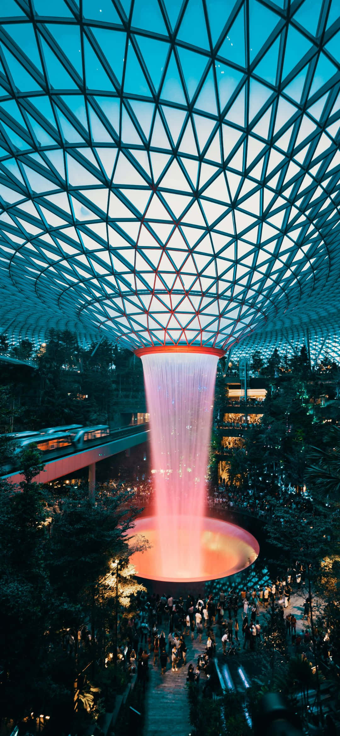 托斯卡尼丘陵的夢幻風景 壁紙 — 一座高聳的室內瀑布在幾何玻璃穹頂下傾瀉而下，人群聚集在下方茂密的熱帶中庭。