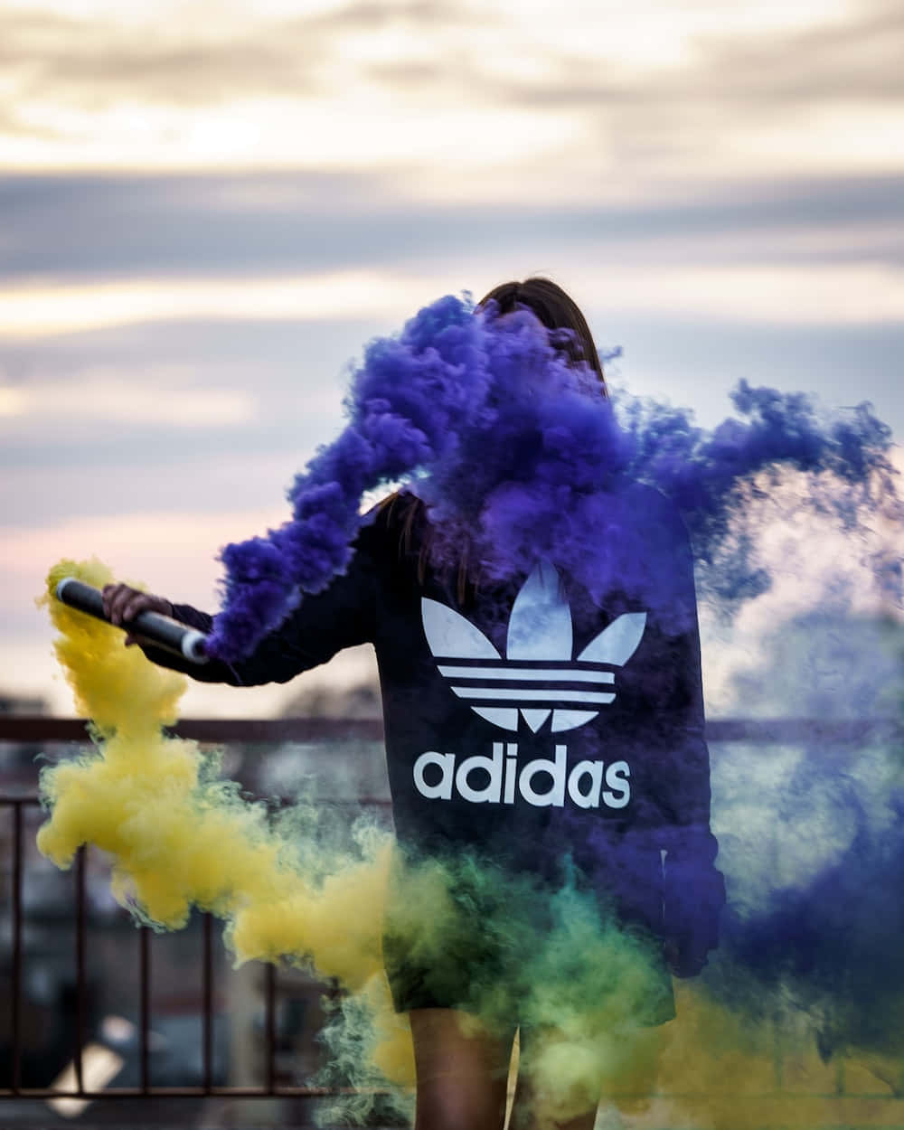 找到你的風格，讓它屬於你 壁紙 — 一個穿著黑色Adidas連帽衫的人站在屋頂上，在柔和的暮色天空中釋放出充滿活力的紫色和黃色煙霧。