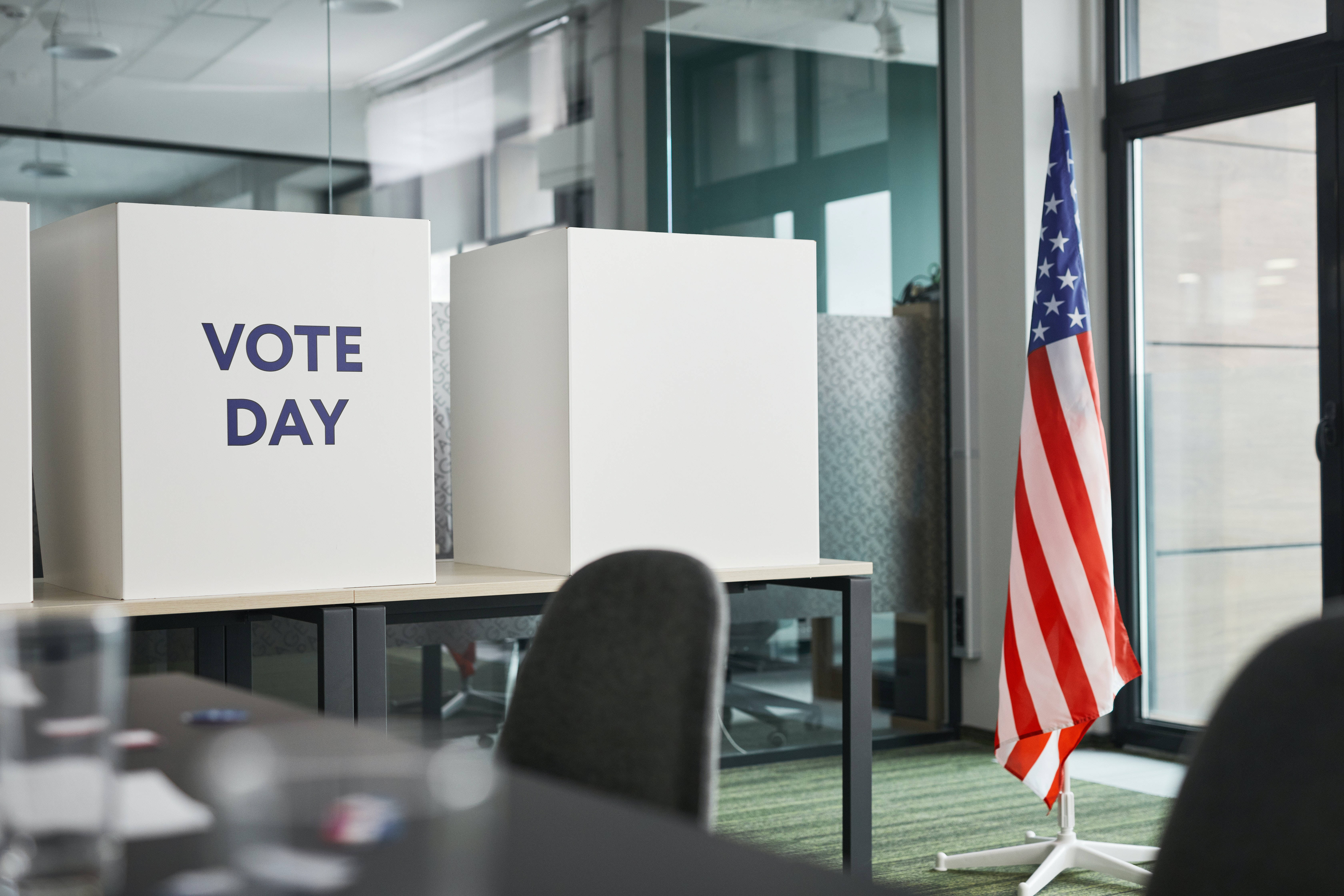投票日的美國國旗 壁紙 — 白色投票亭帶有「VOTE DAY」文字，位於現代辦公空間，旁邊有美國國旗。