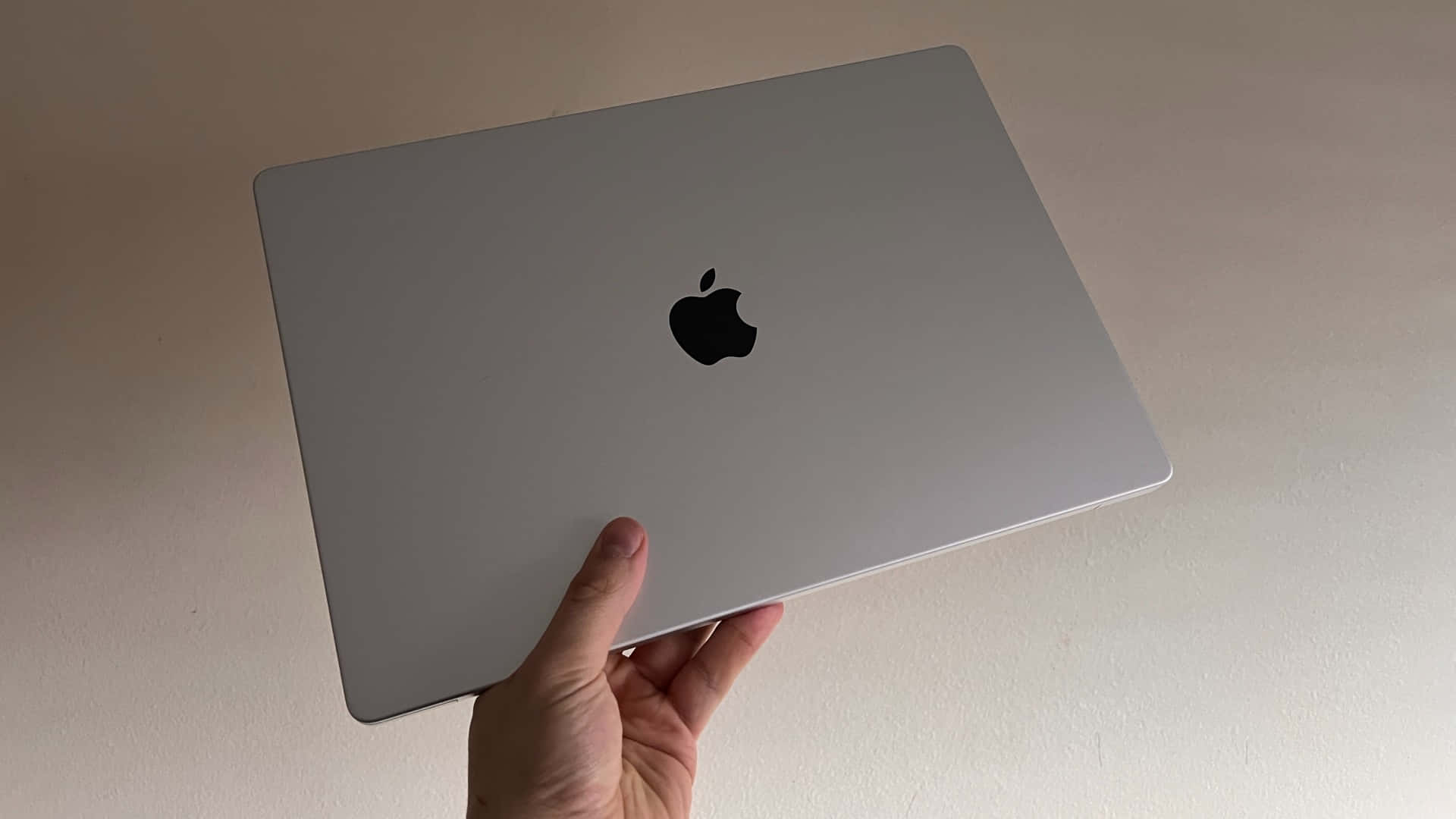 投資 MacBook 的強大效能 壁紙 — 一隻手拿著一部銀色 MacBook 手提電腦，帶有黑色 Apple 標誌，背景是純色米白色牆壁。