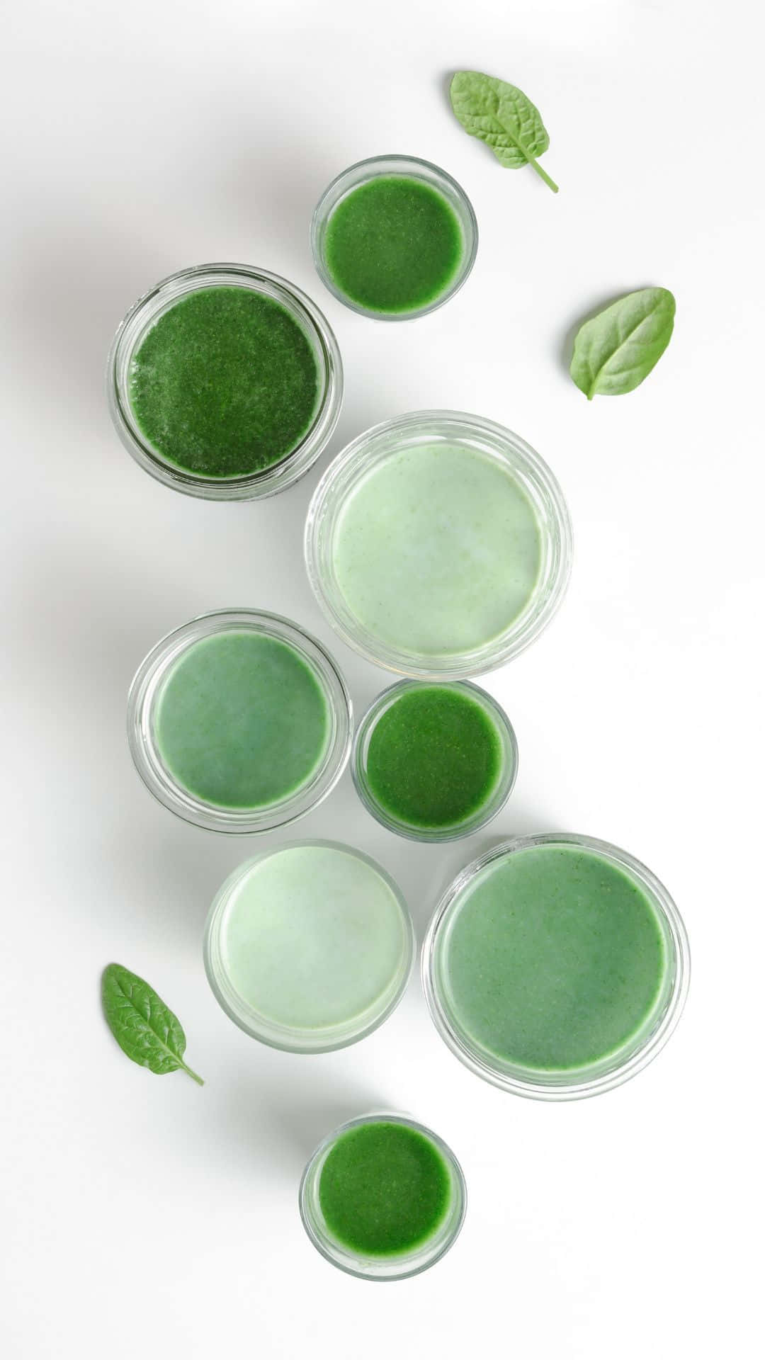 抹茶綠茶 sage 風格 壁紙 — 從上方俯瞰，玻璃罐中裝有各種綠色冰沙，散落著新鮮菠菜葉，置於明亮的白色表面上。